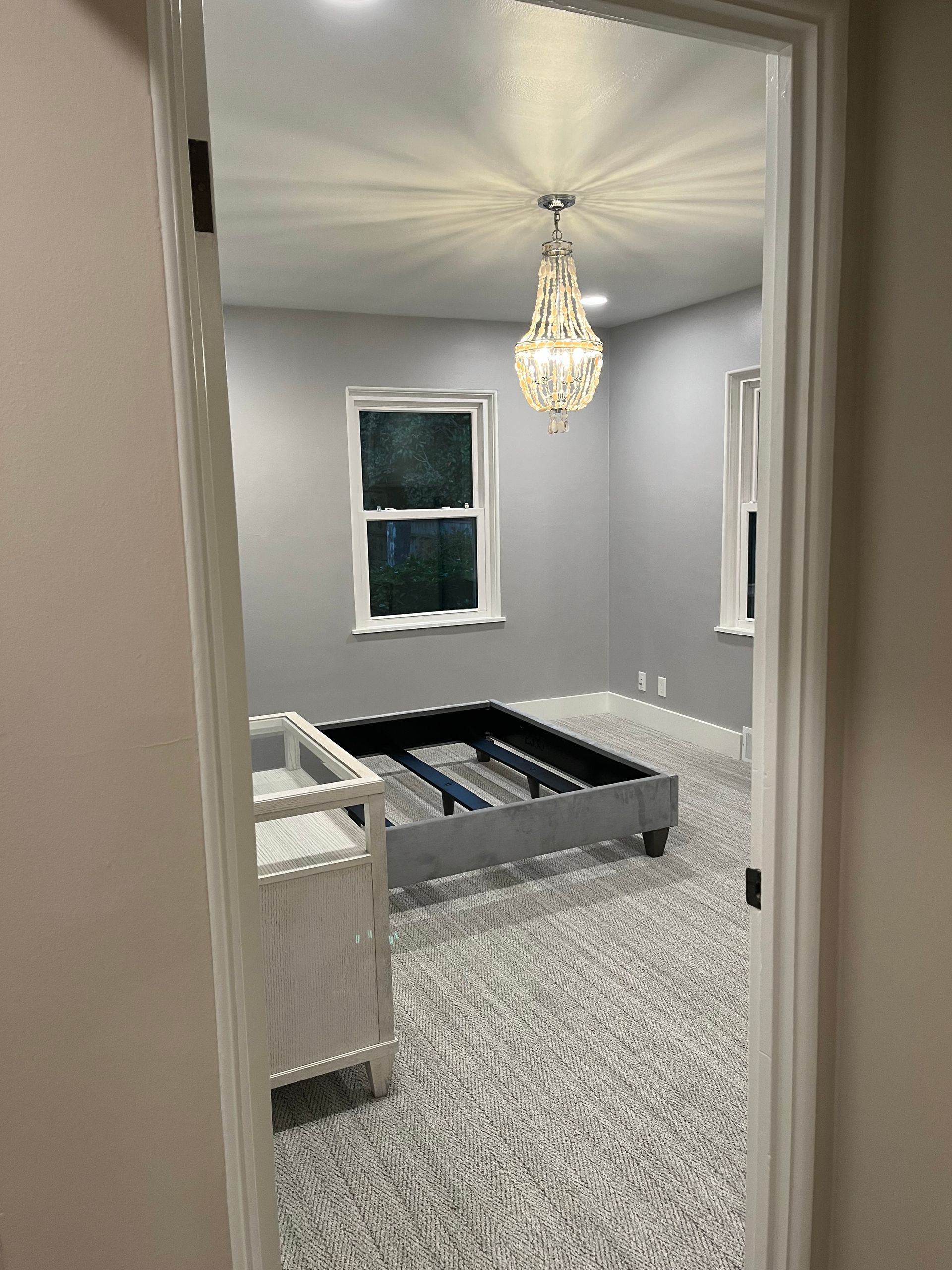 Unfinished bedroom with gray carpet, crib frame, white walls, and a chandelier hanging from the ceiling