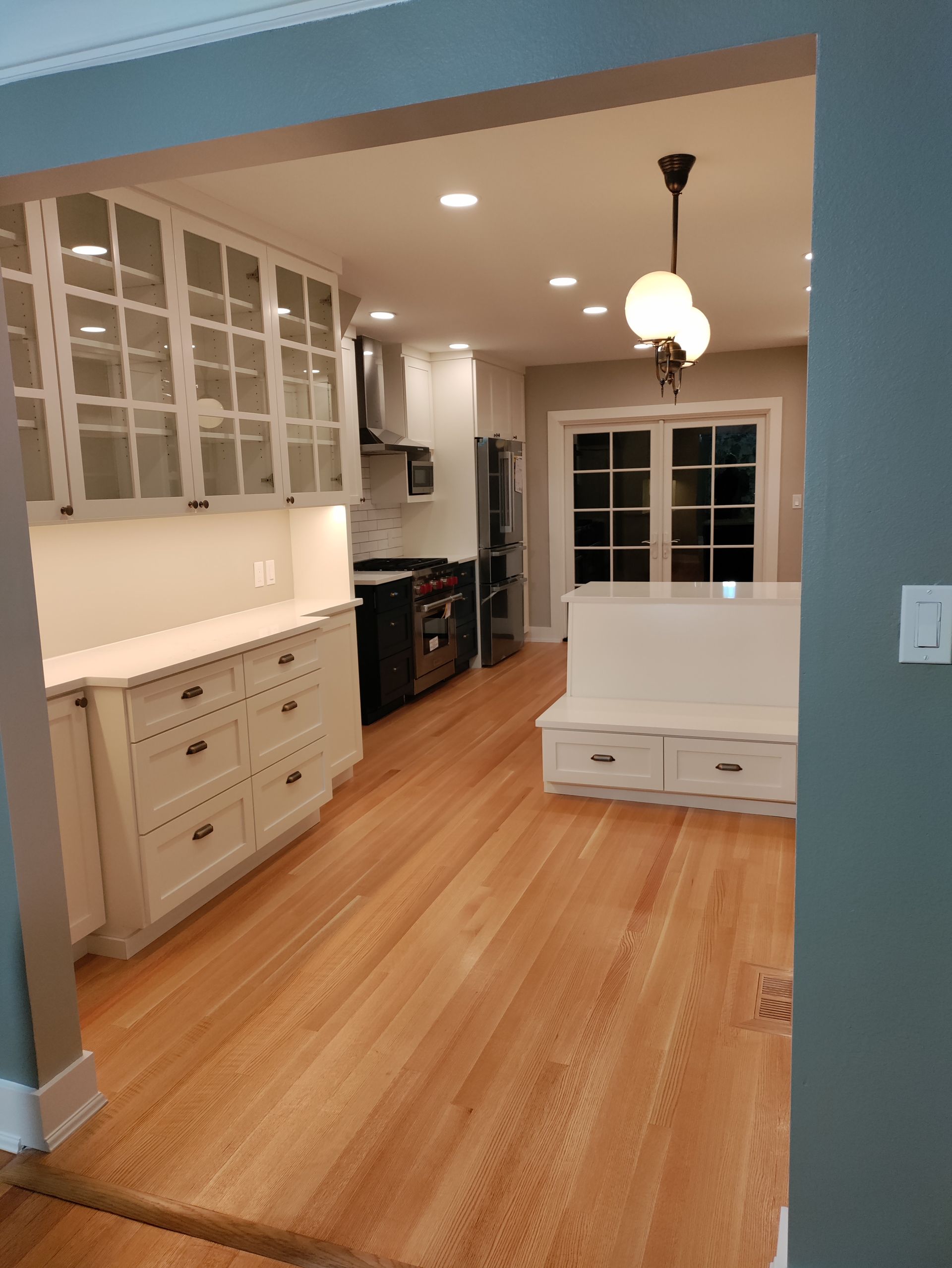 Bright empty kitchen with light wood floors, white cabinets, and a small bench by a window.