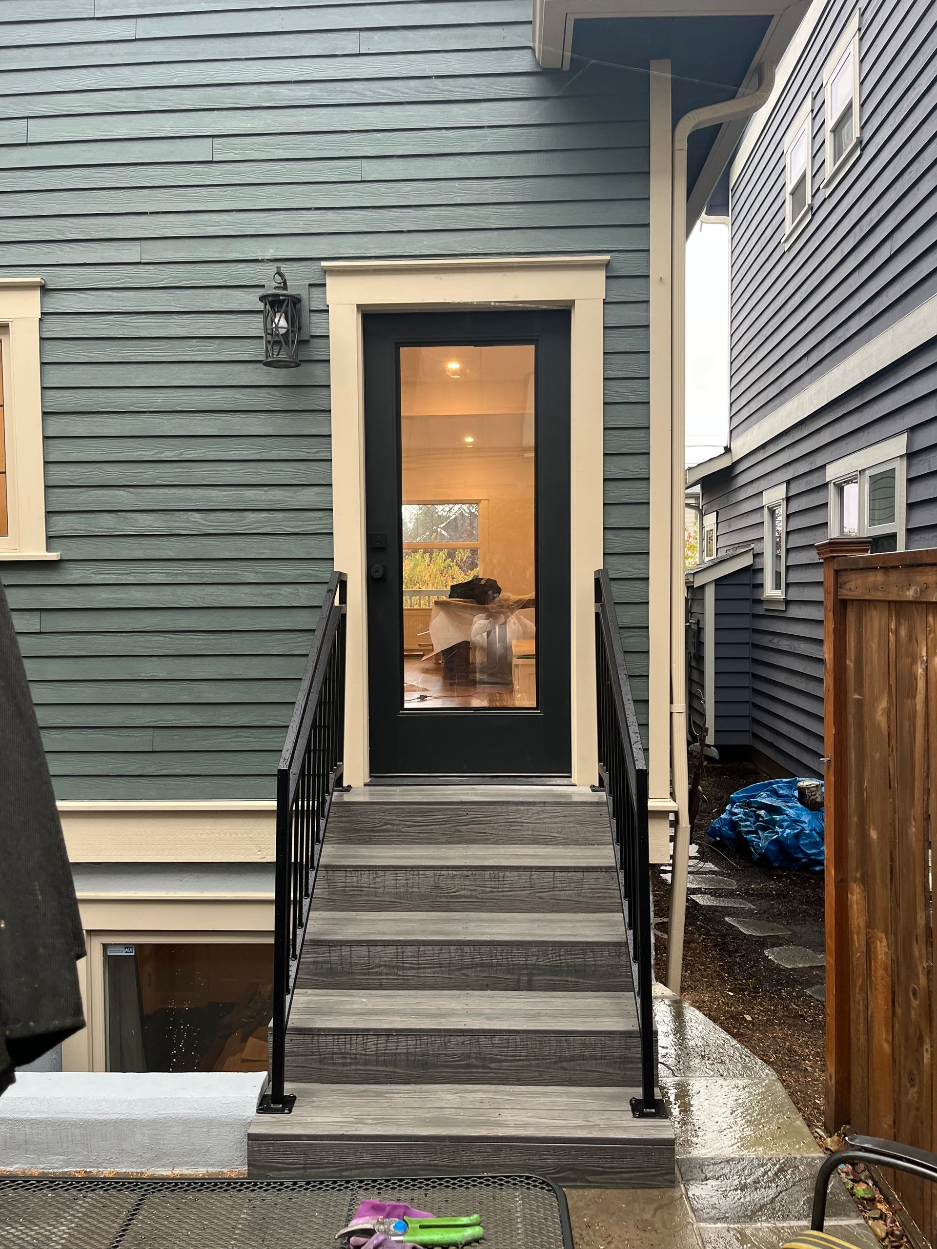 Back door with steps and black railing on a green house, seen in a narrow outdoor passage.