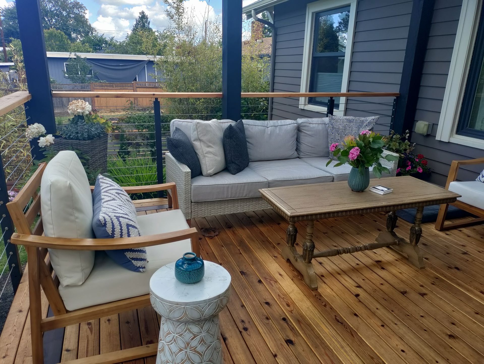 Wooden patio with cushioned outdoor seating, coffee table, and flowers beside a house.