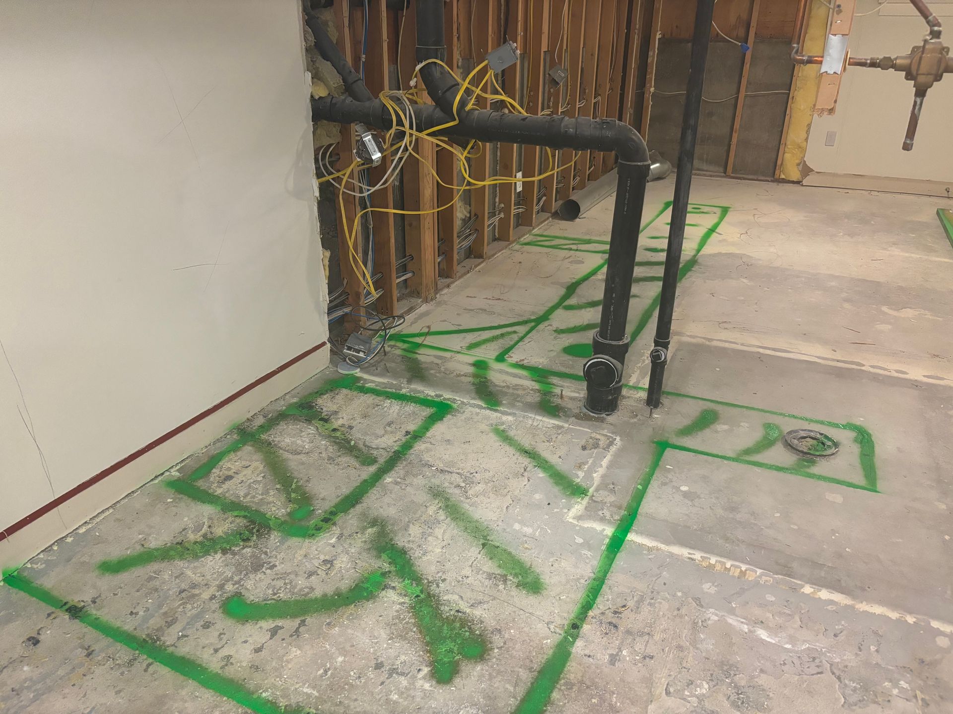 Green tape markings on a concrete floor around exposed pipes and a wall, in a construction area