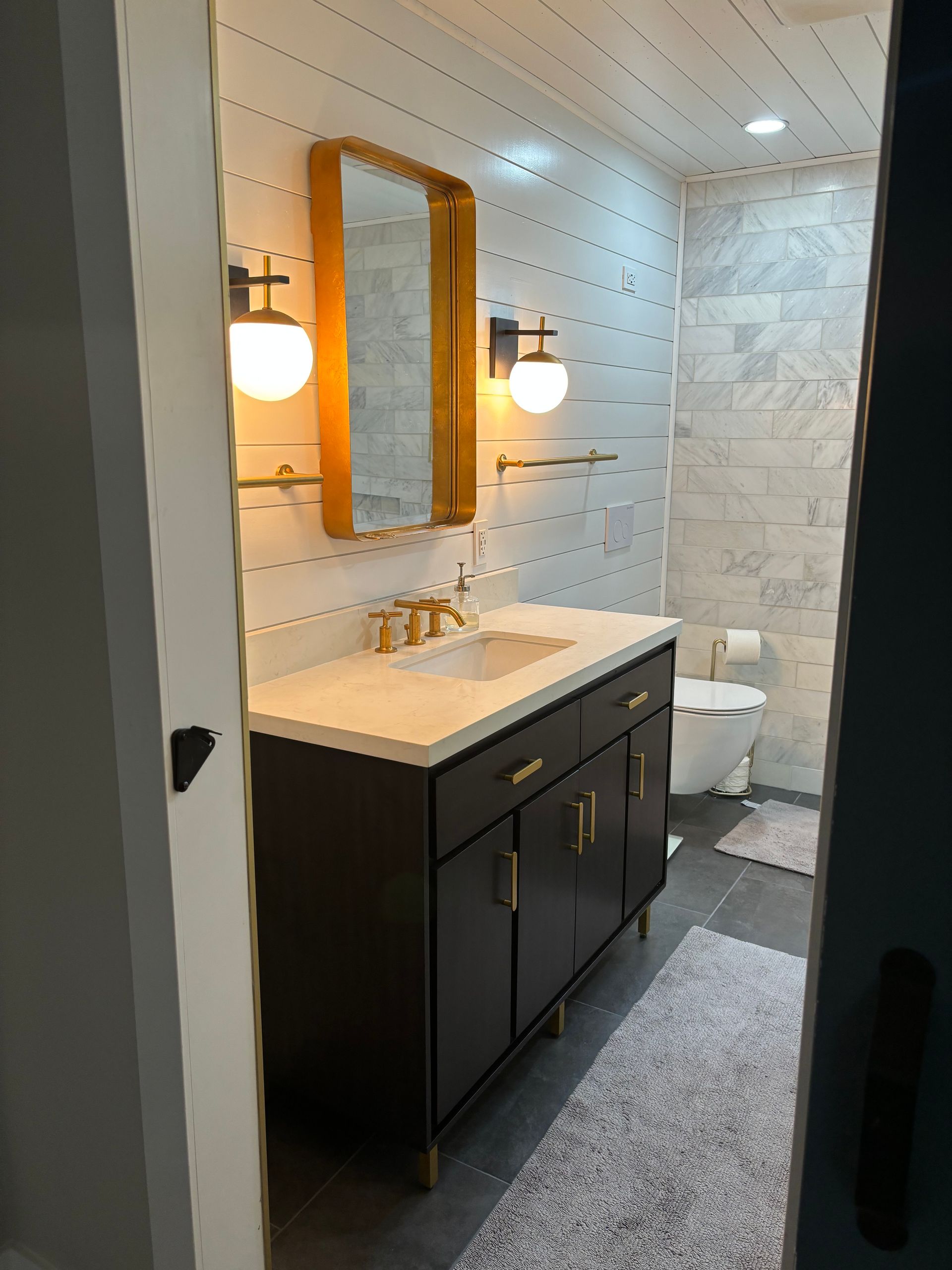 Modern bathroom with dark vanity, white sink, gold mirror, wall sconces, and tiled shower.