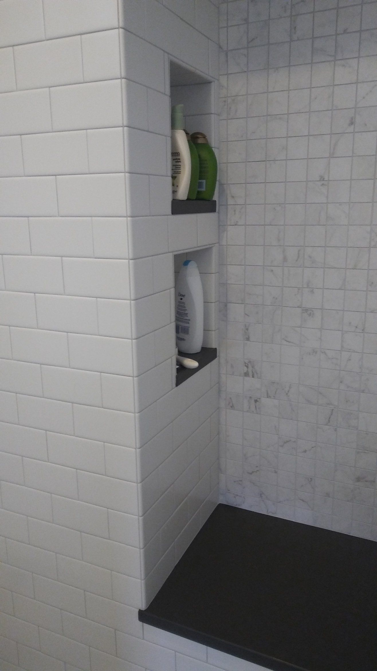 White tiled shower niche with green and white bottles on shelves in a bathroom.