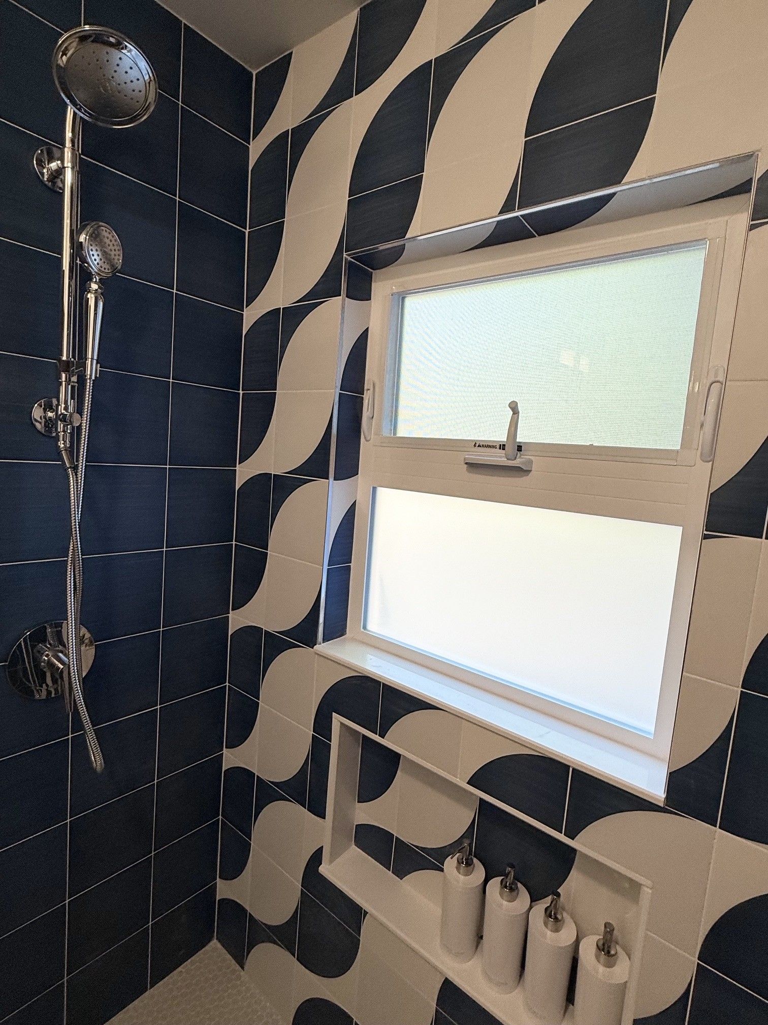 Black-and-white wavy tiled bathroom with showerhead, window, and shelf with toiletries.