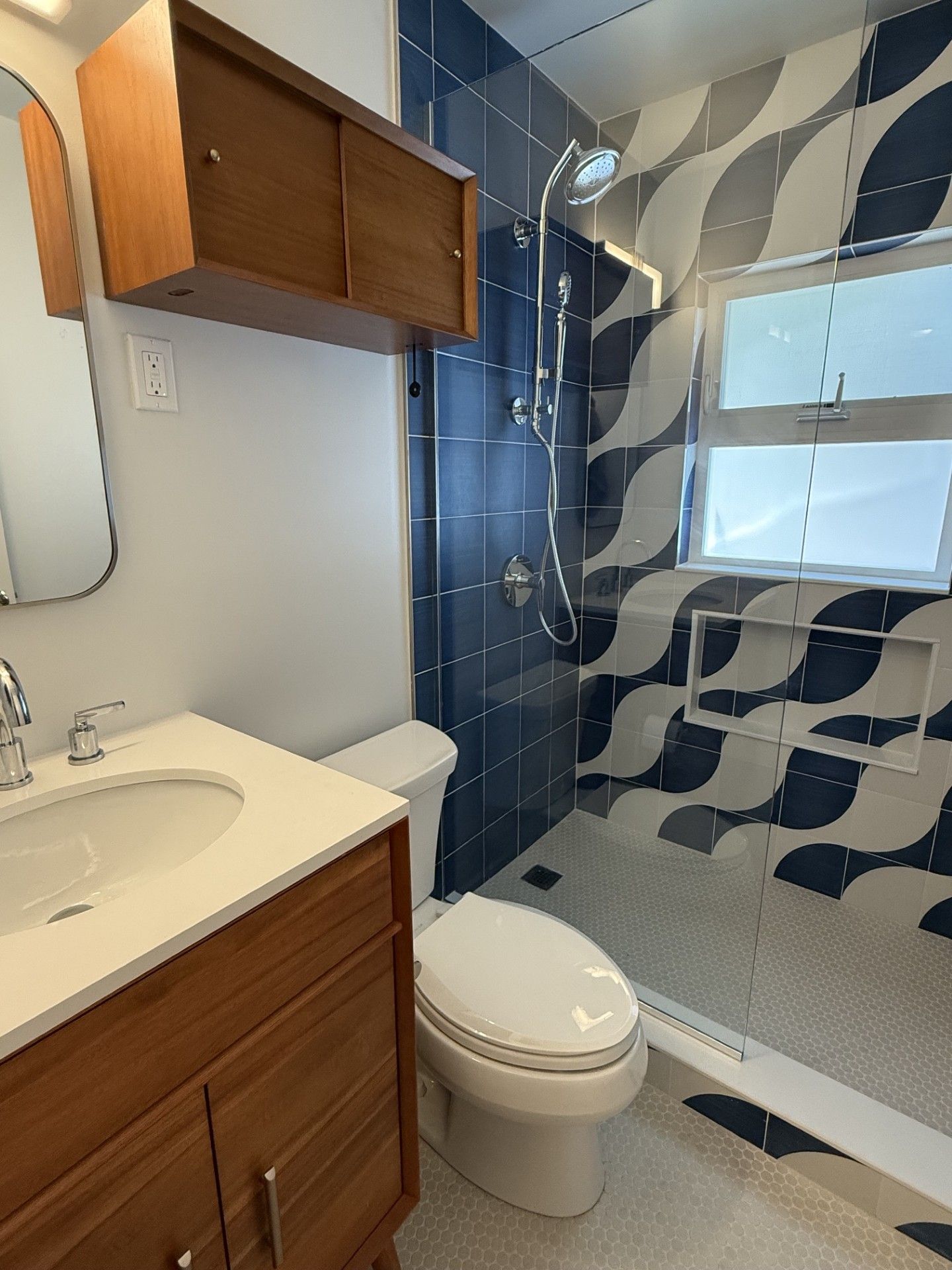 Small bathroom with wood vanity, white toilet, blue tile shower, and patterned shower curtain.