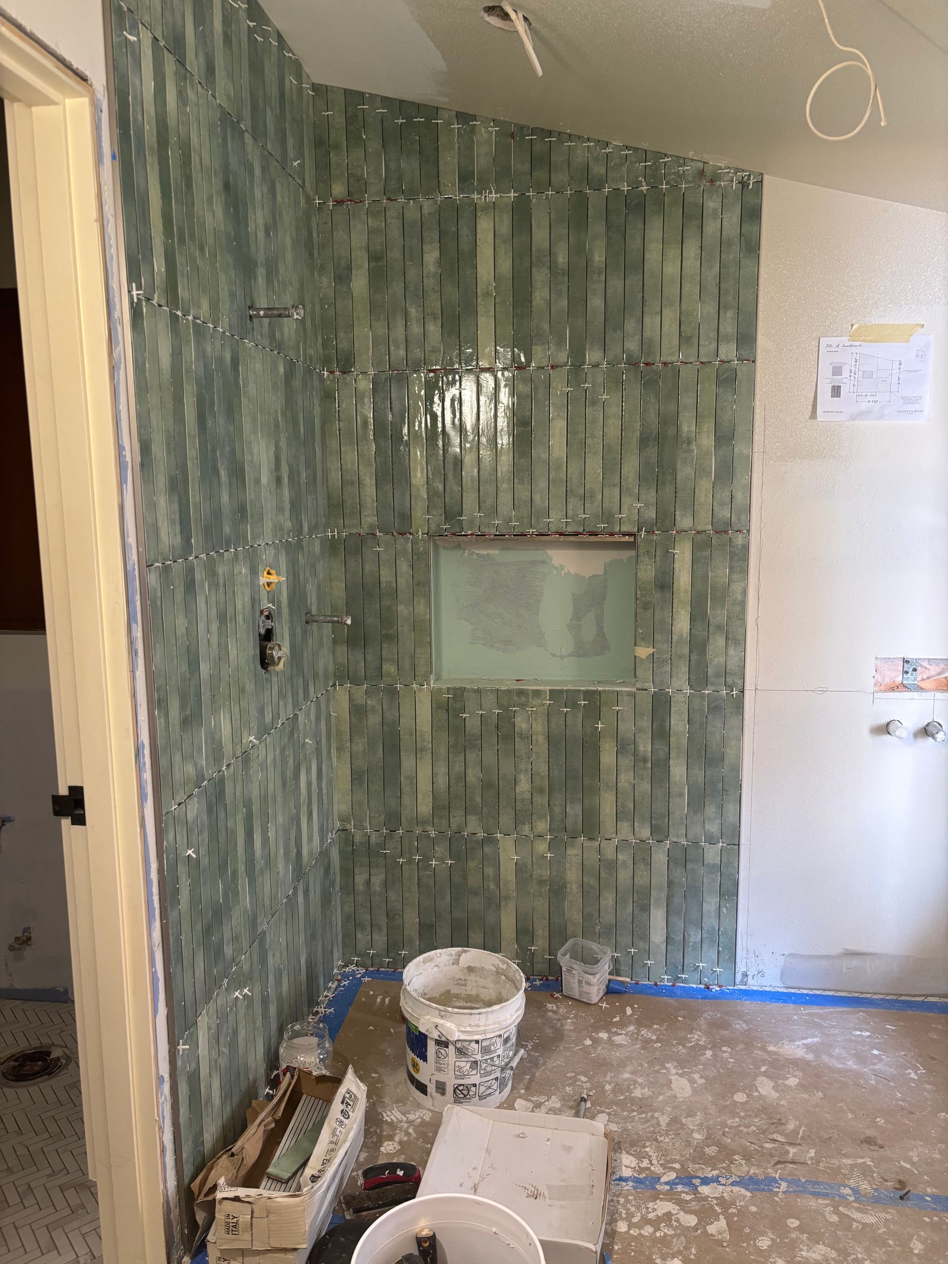 Bathroom shower wall under renovation, covered in green tile with construction materials on the floor.