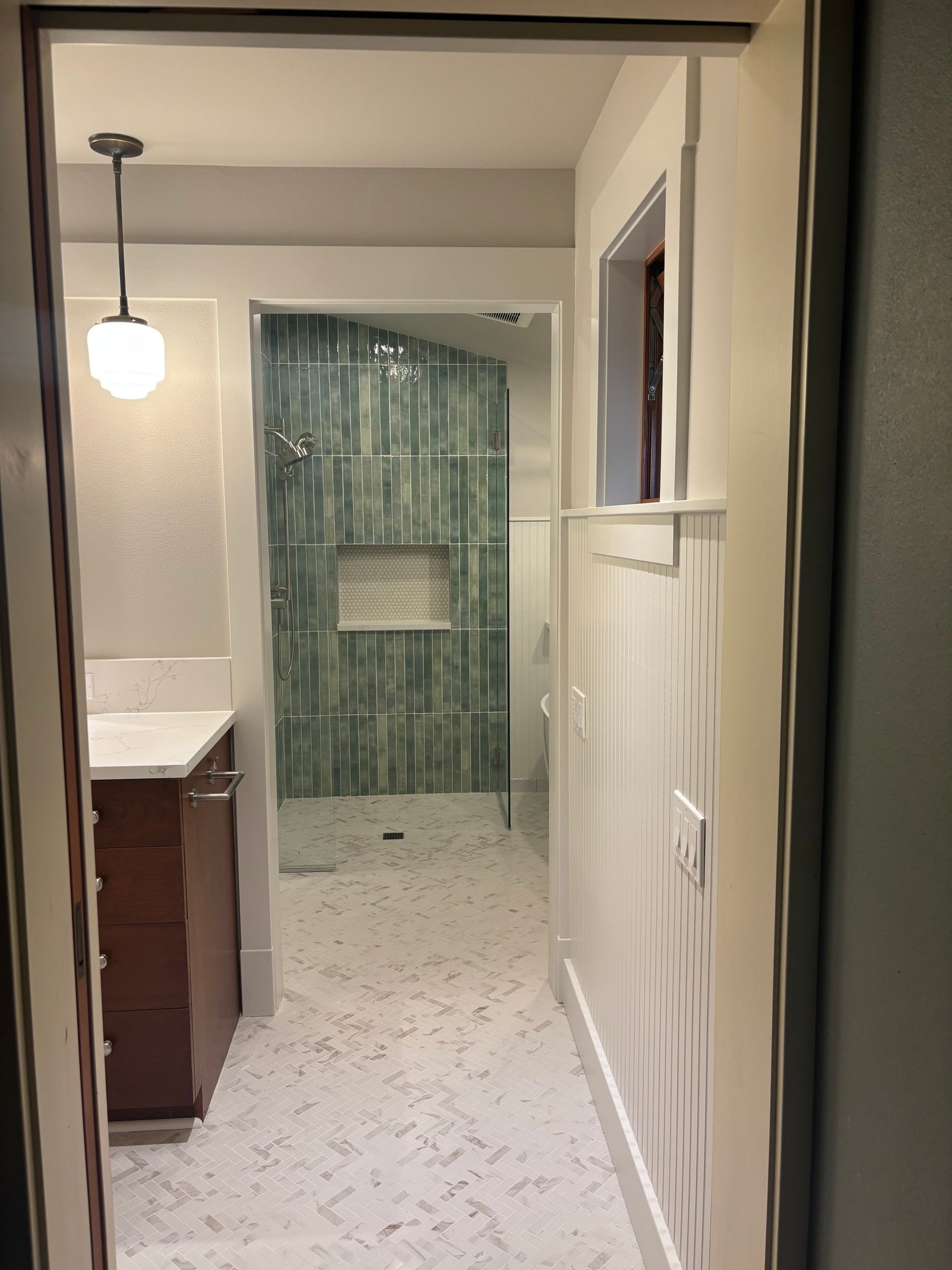 Narrow hallway with tiled floor, hanging light, vanity on left, and green-tiled bathroom at the end.