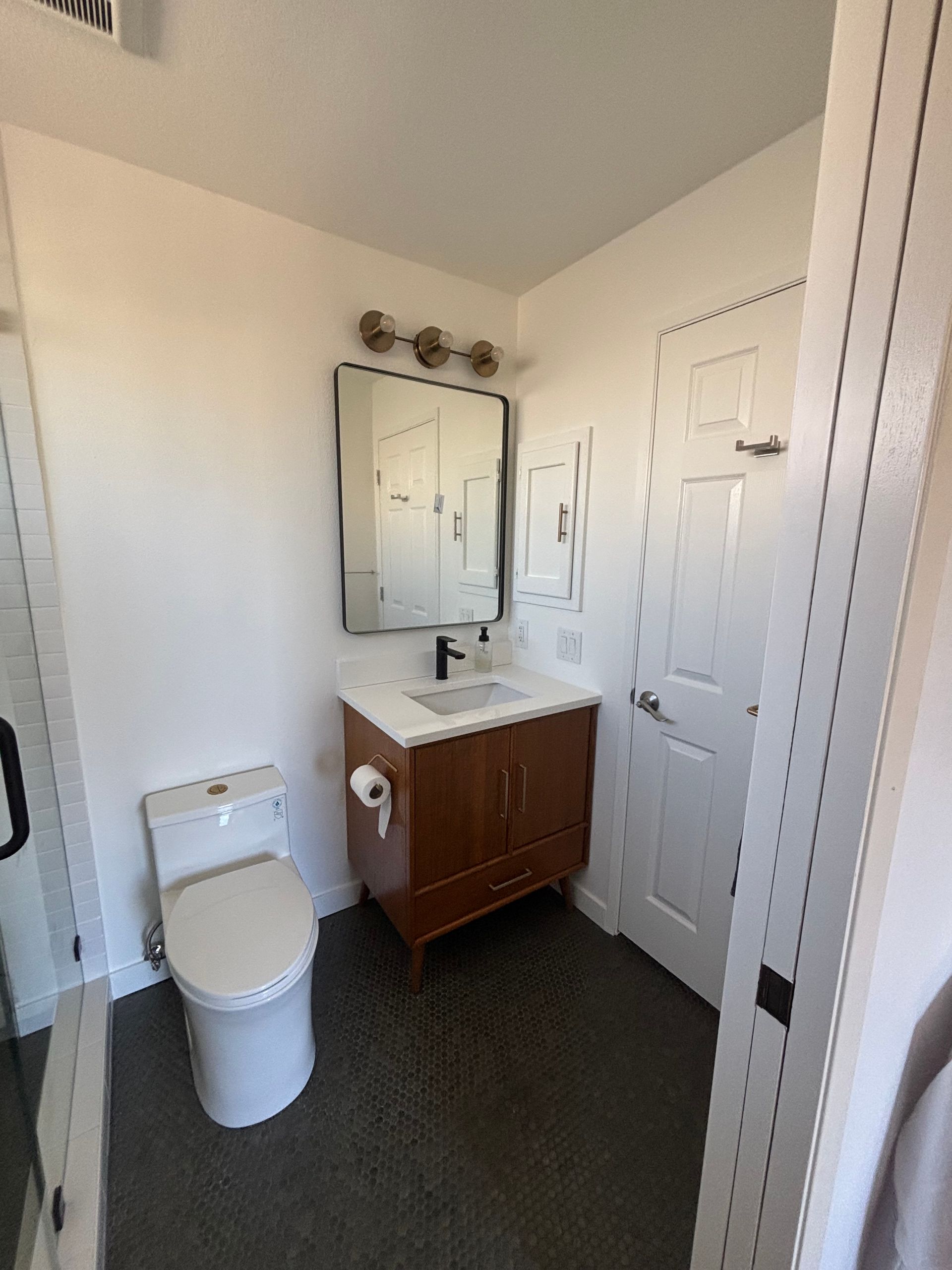 Small bathroom with toilet, wood vanity, mirror, sink, and white paneled door.