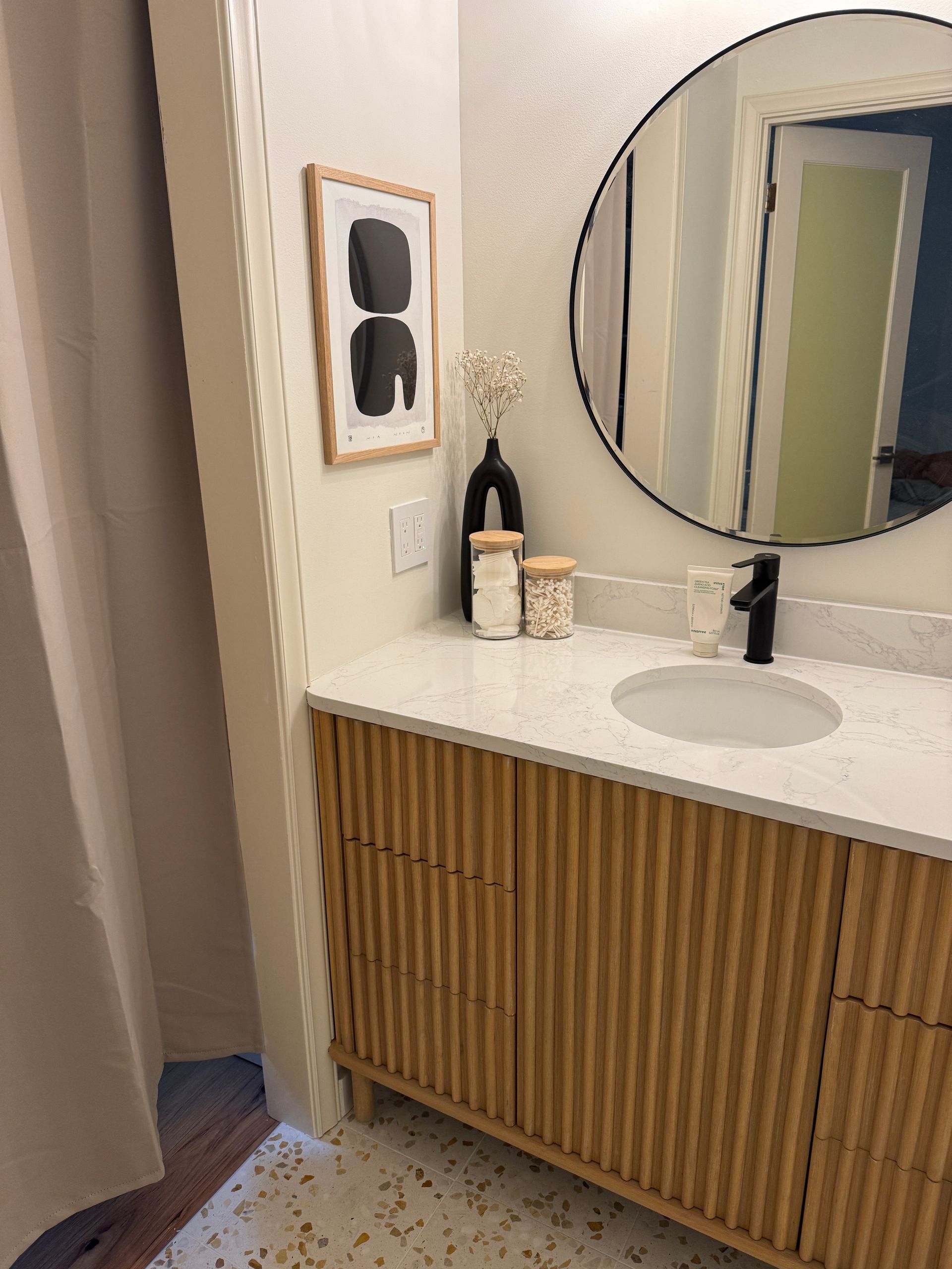 Modern bathroom vanity with wood slatted cabinet, round mirror, white countertop, and black faucet.