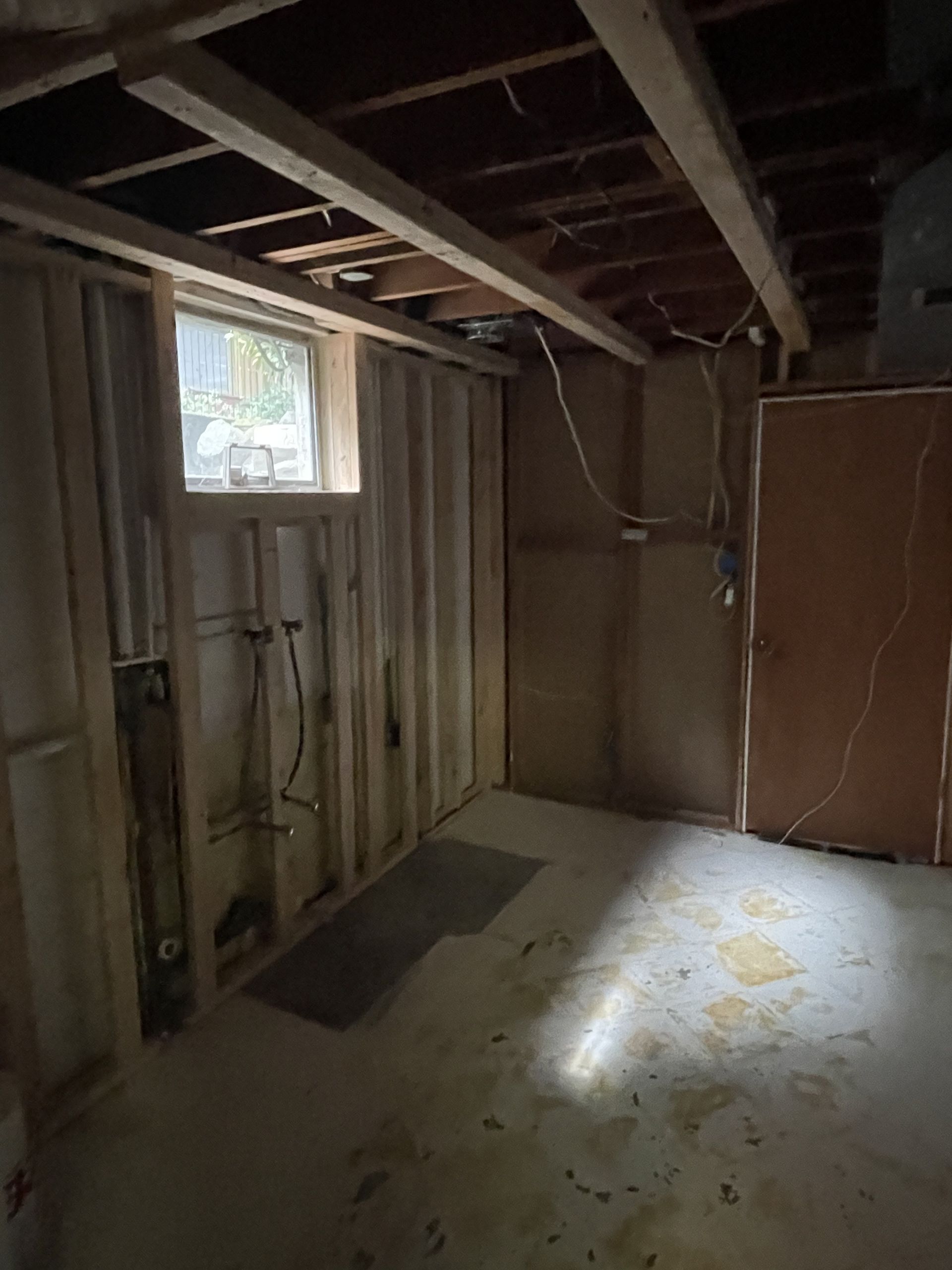 Unfinished basement room with exposed studs, a small window, and light on the concrete floor.