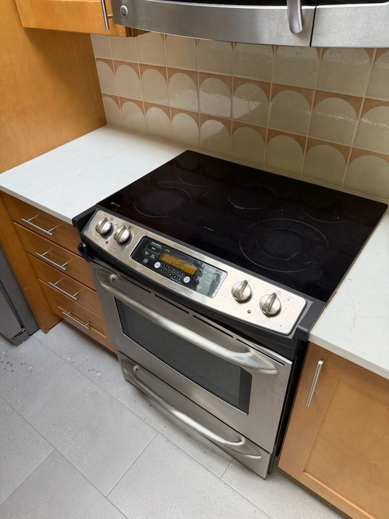 Kitchen stove and oven between wooden cabinets, with black glass cooktop and patterned tile backsplash.