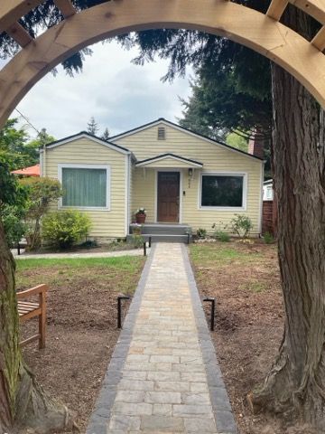 Small yellow house with a stone walkway, viewed through an arched wooden frame and trees.