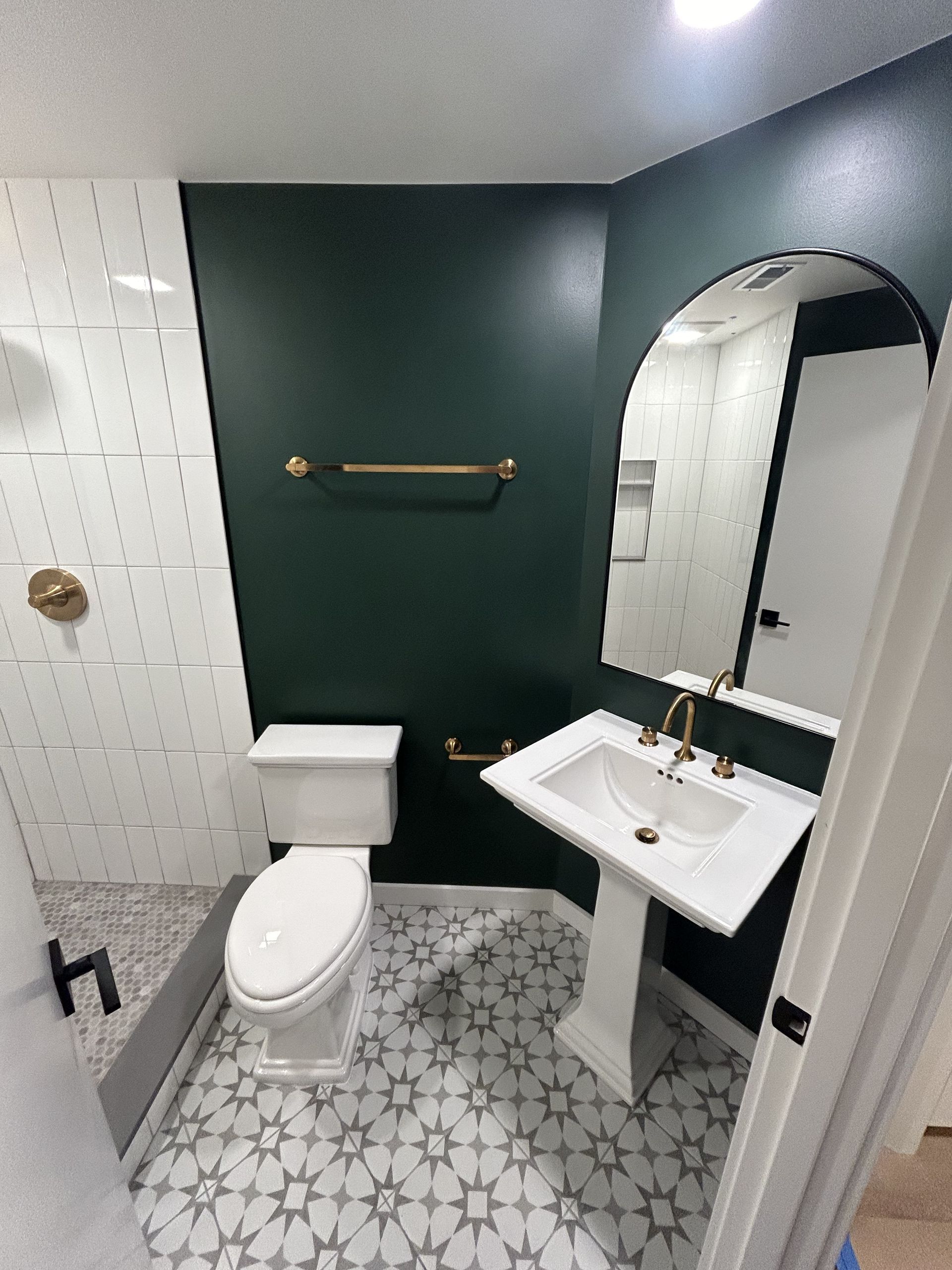 Small bathroom with toilet, pedestal sink, round mirror, dark green accent wall, and patterned tile floor.