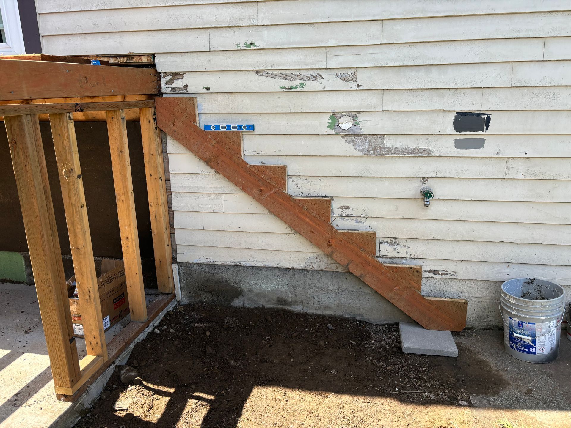 Outdoor wooden staircase beside a white house wall, with a bucket on the ground and deck railing on the left.