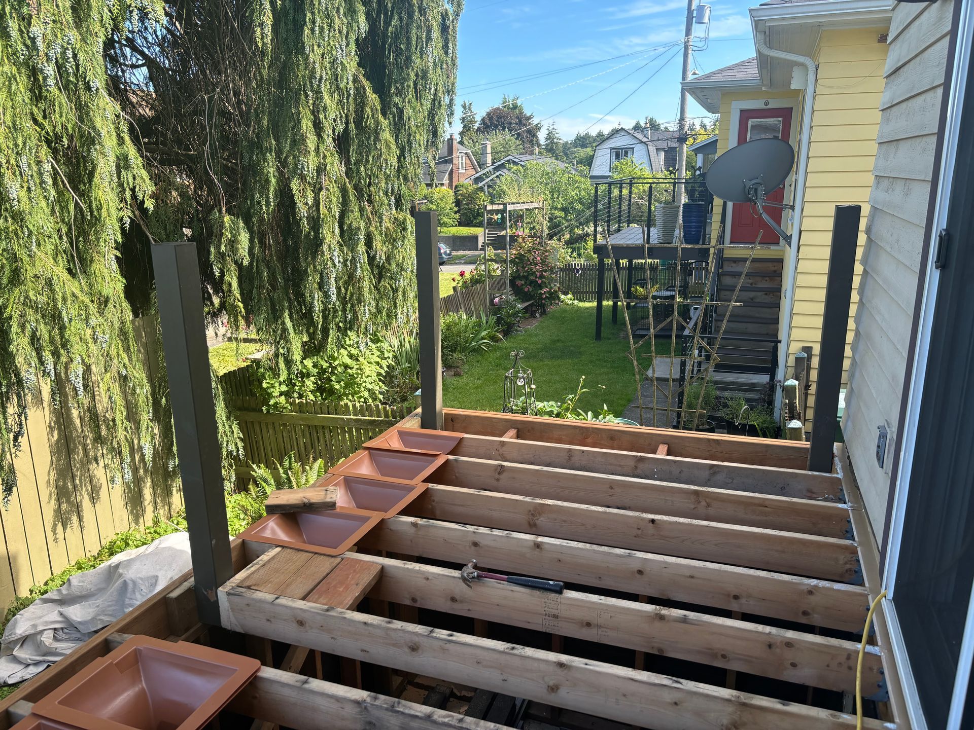 Backyard deck under construction beside a yellow house, with exposed beams and lumber stacked nearby