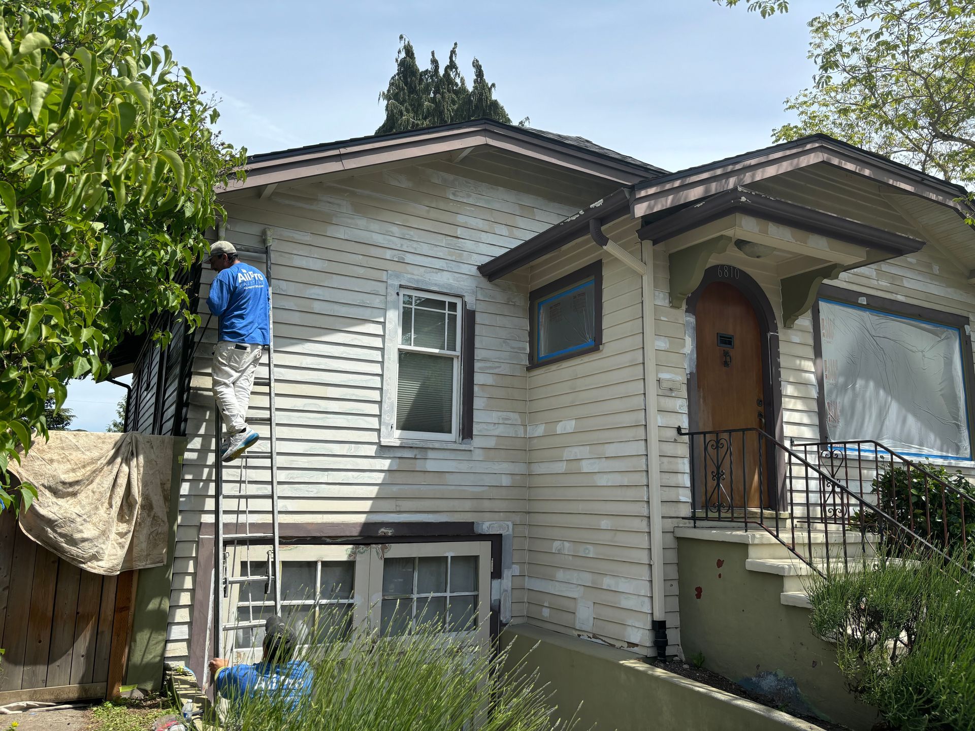 Worker on a ladder painting the exterior of a beige house beside a front porch