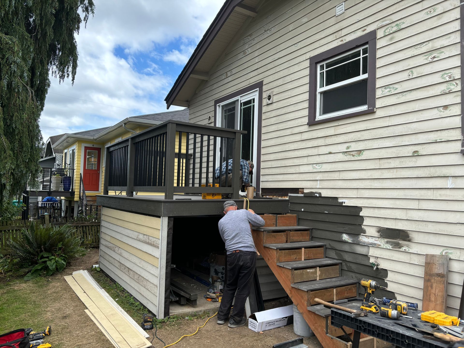 Person building a wooden deck staircase beside a light-gray house with black railings