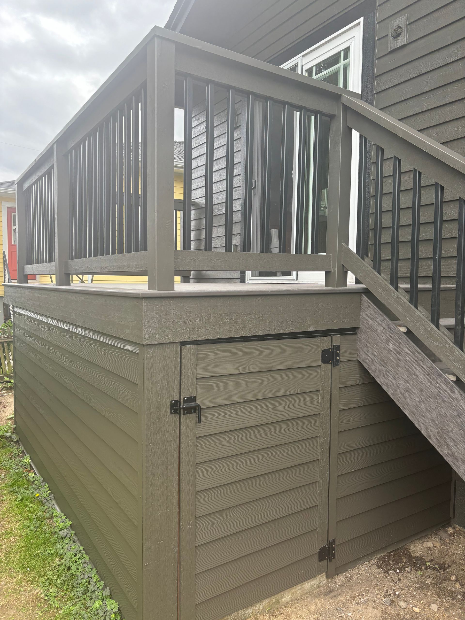 Raised deck with gray railings and stairs over a brown storage area beside a house.