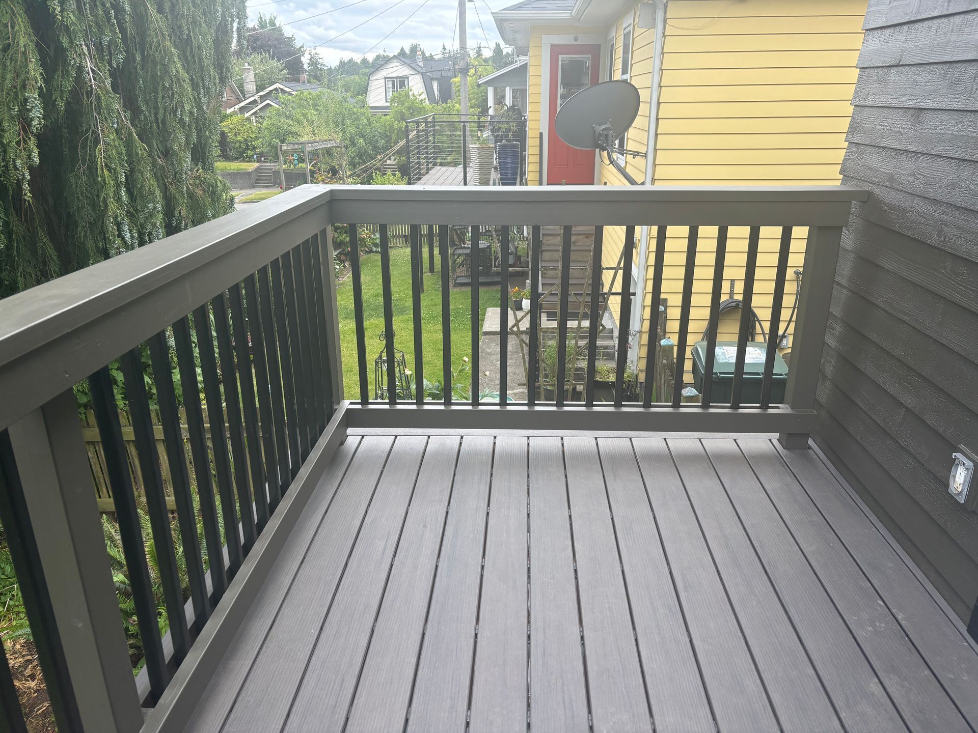 Small balcony with dark railings, gray floorboards, and a view of a yellow house and trees.