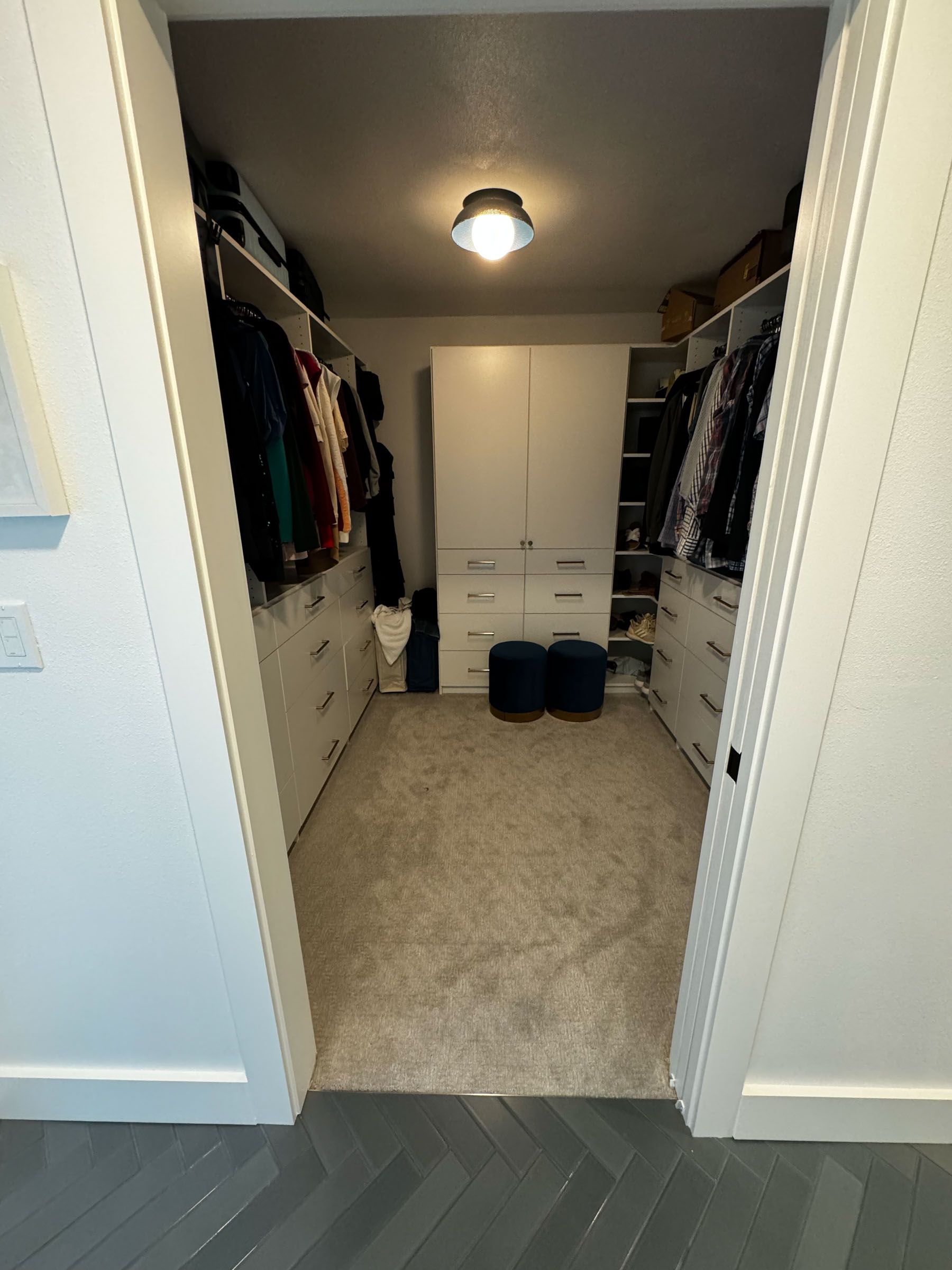 Open walk-in closet with hanging clothes, white shelving, and a ceiling light above beige carpet.