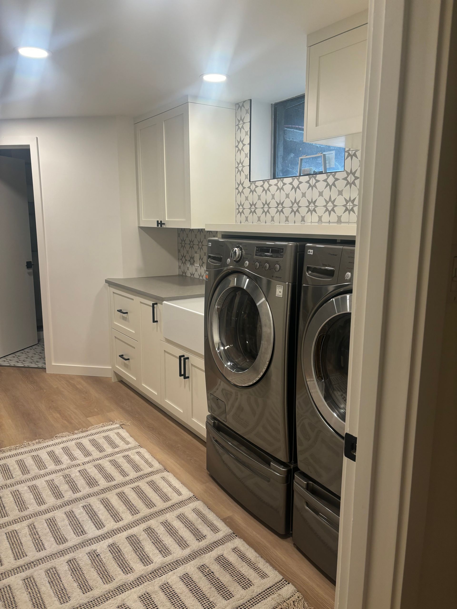 Laundry room with white cabinets, patterned backsplash, and a pair of dark front-loading washer-dryers.