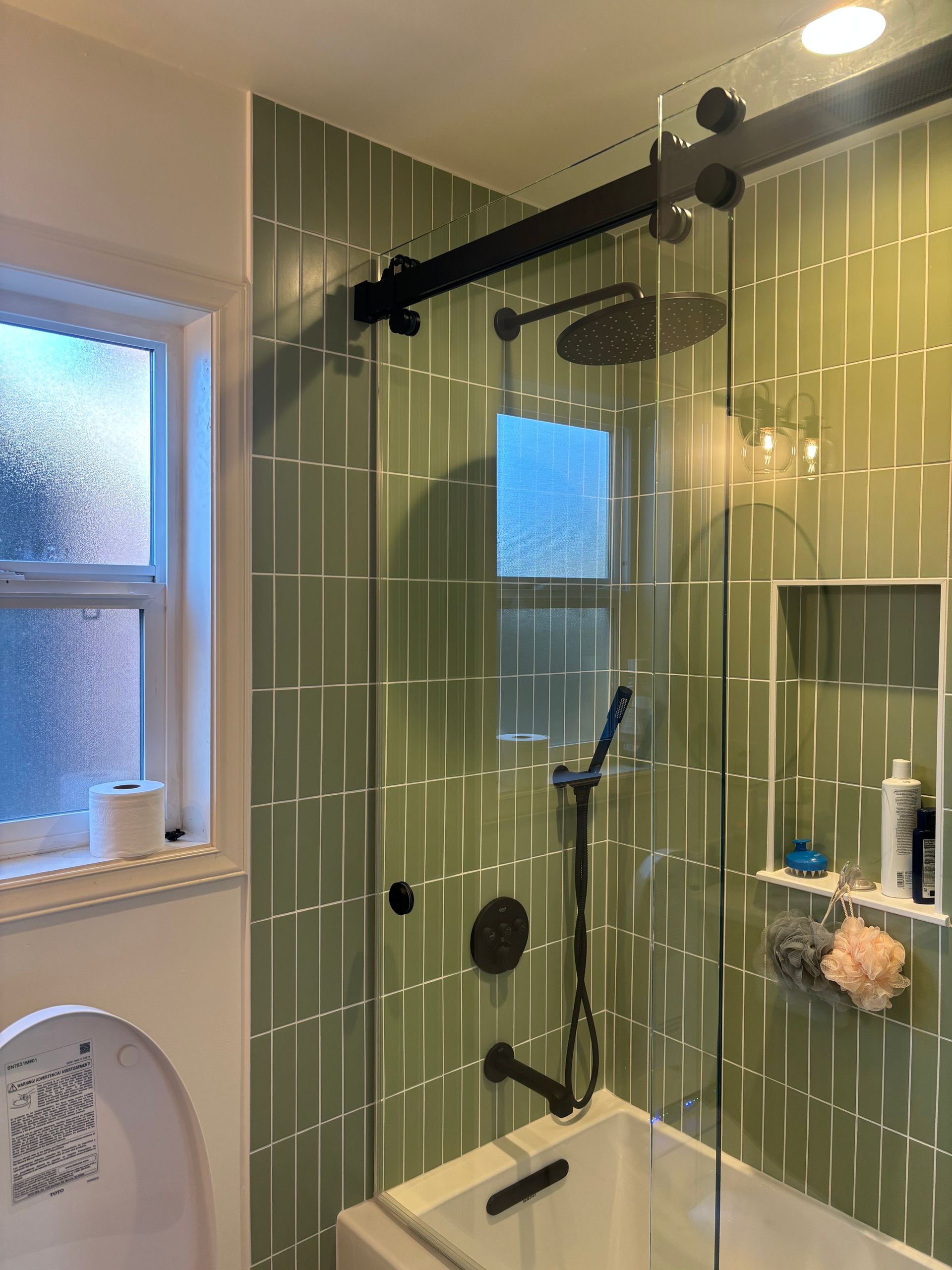 Green-tiled bathroom with a glass shower, window, toilet, and recessed shelving.