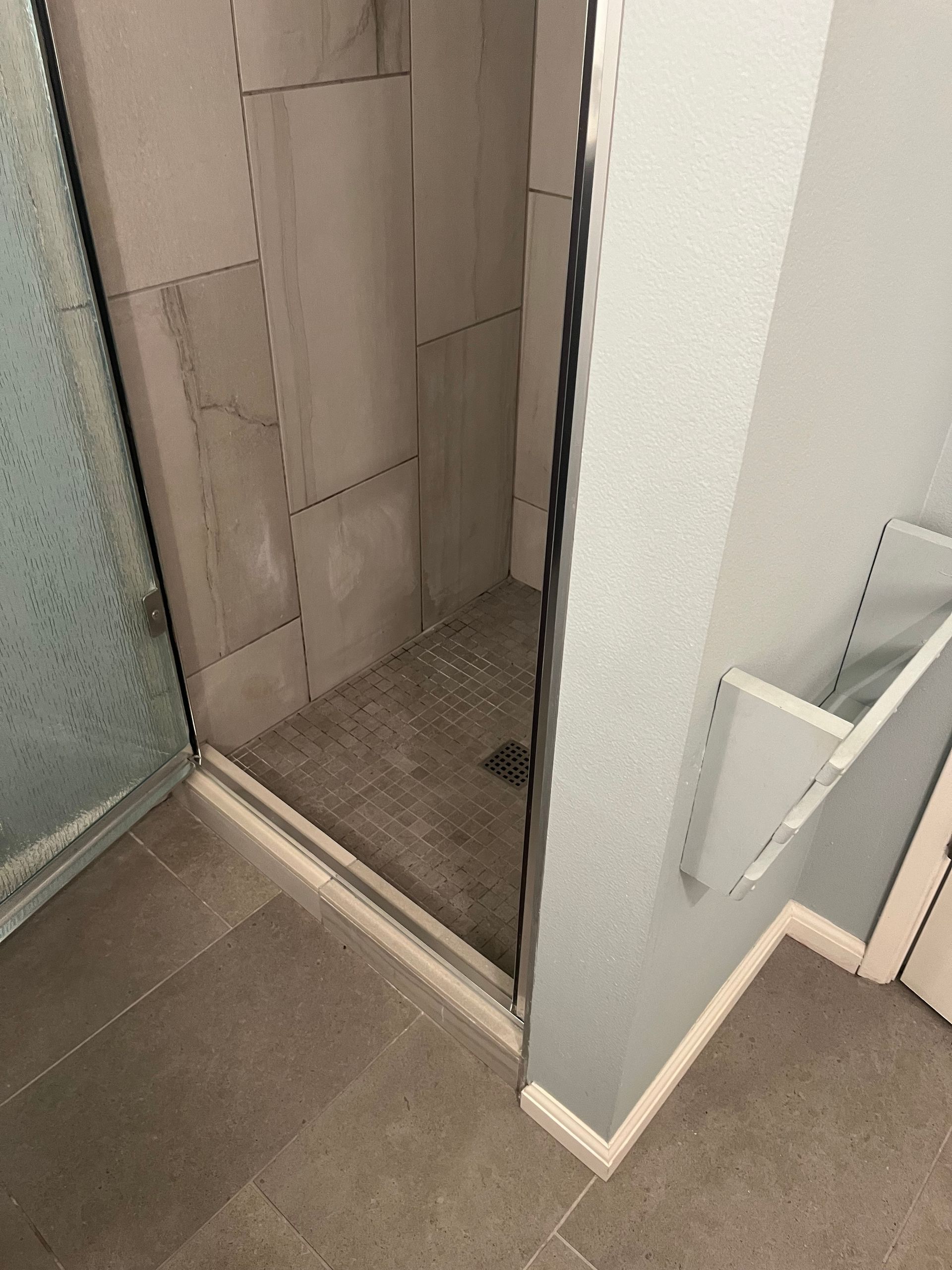 Open shower stall with gray tiled walls, frosted glass door, and beige bathroom floor.