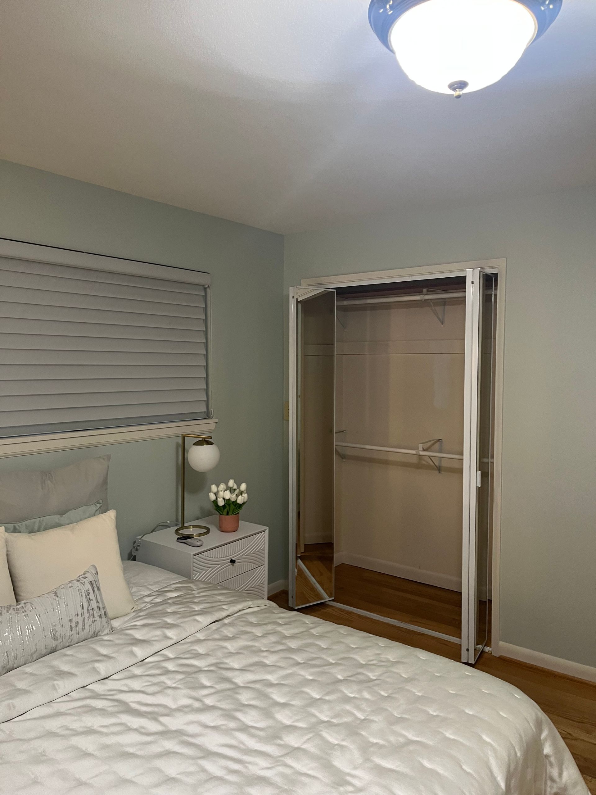 Cozy bedroom with a white bed, nightstand, window blinds, and open closet doors.