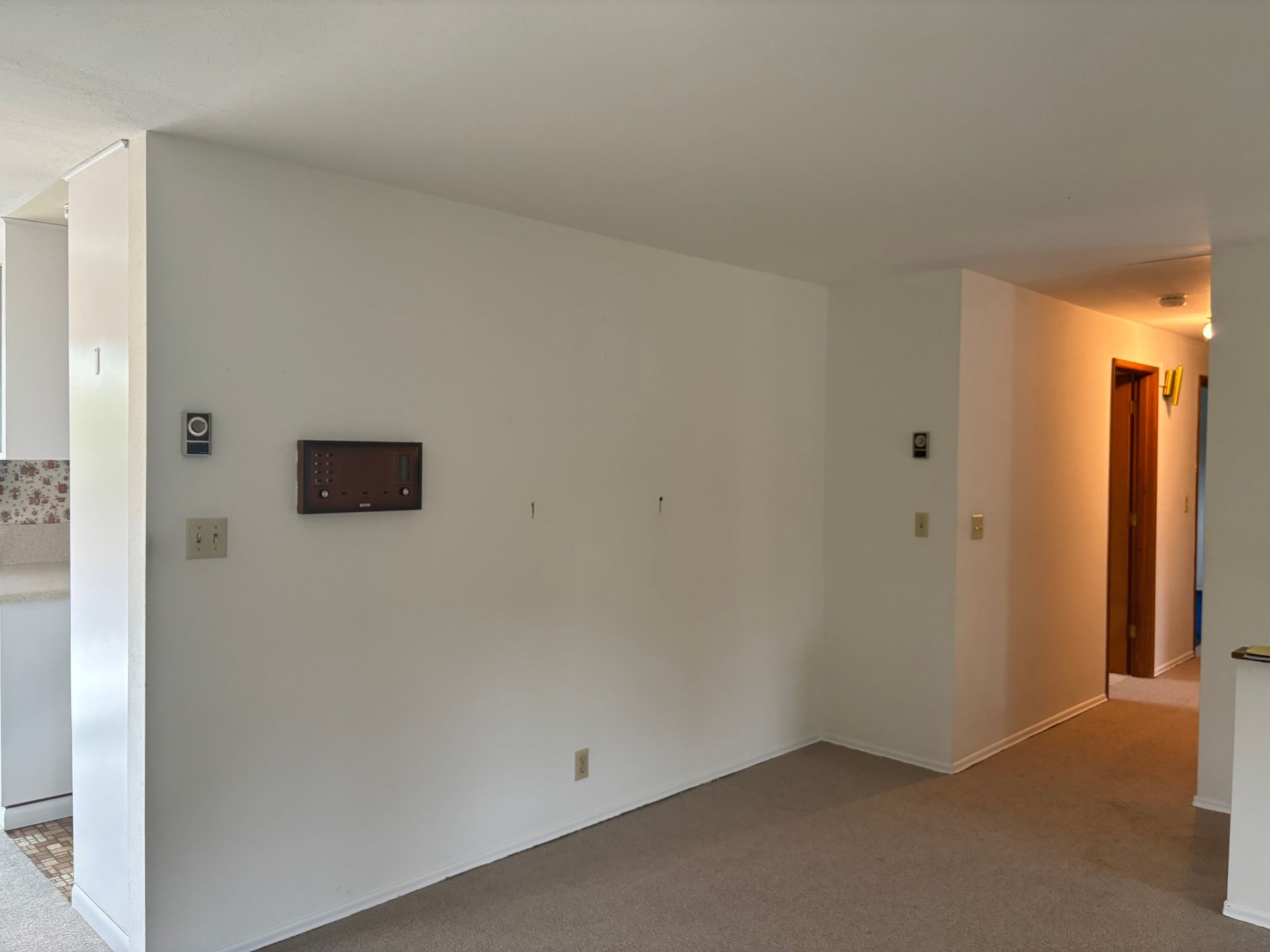 Empty beige living room with carpet, white walls, and a lit hallway opening to the right