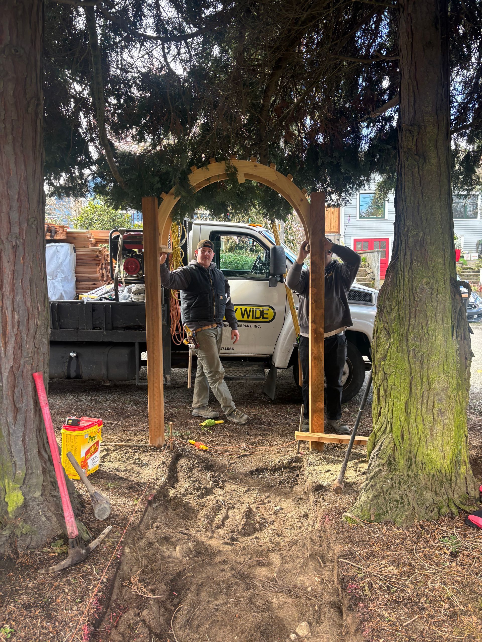 Worker standing under a wooden arch in a wooded yard, with a truck and construction materials behind him.