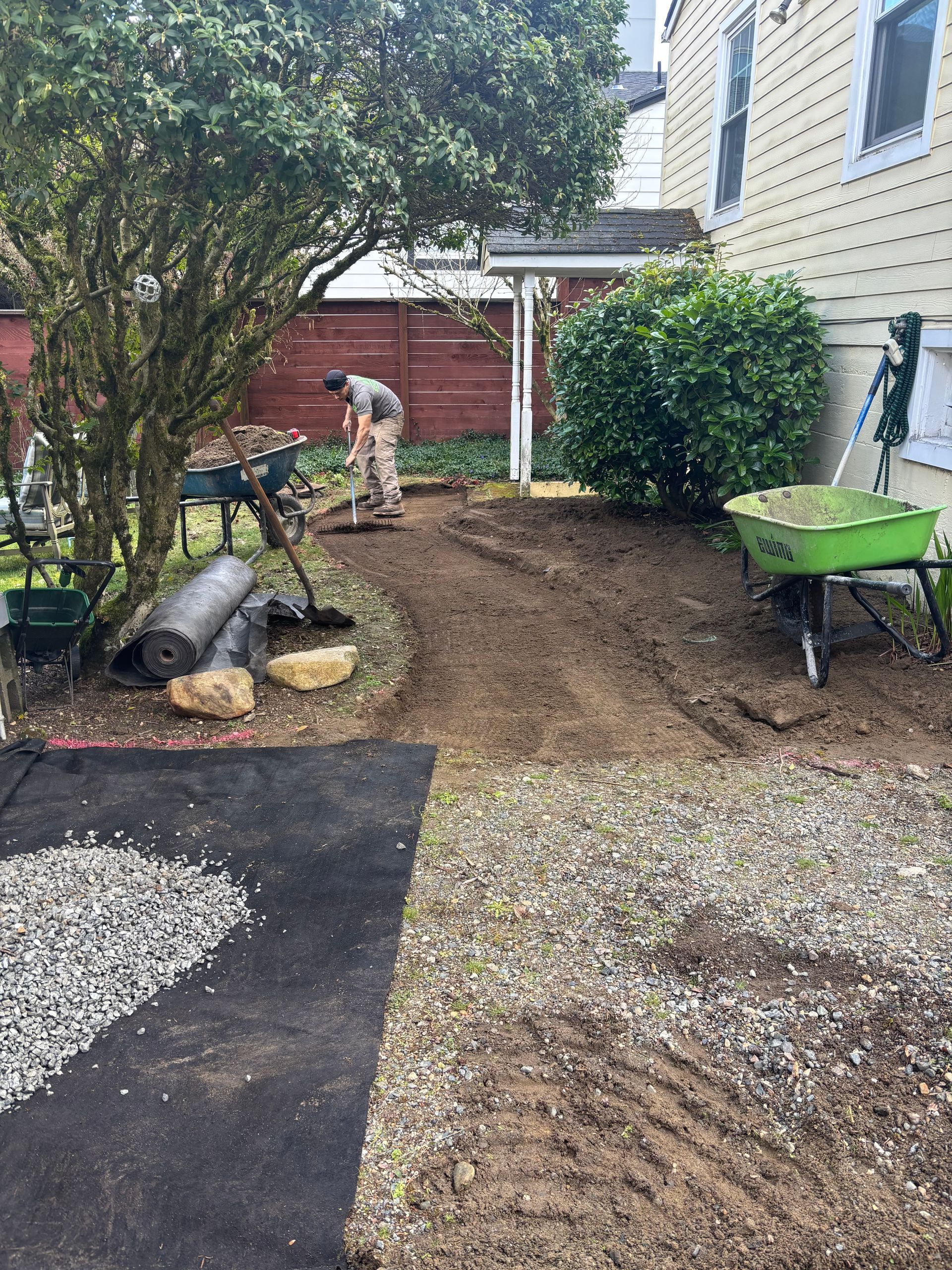 Person digging soil in a backyard beside a house, with a wheelbarrow and fresh gravel path.