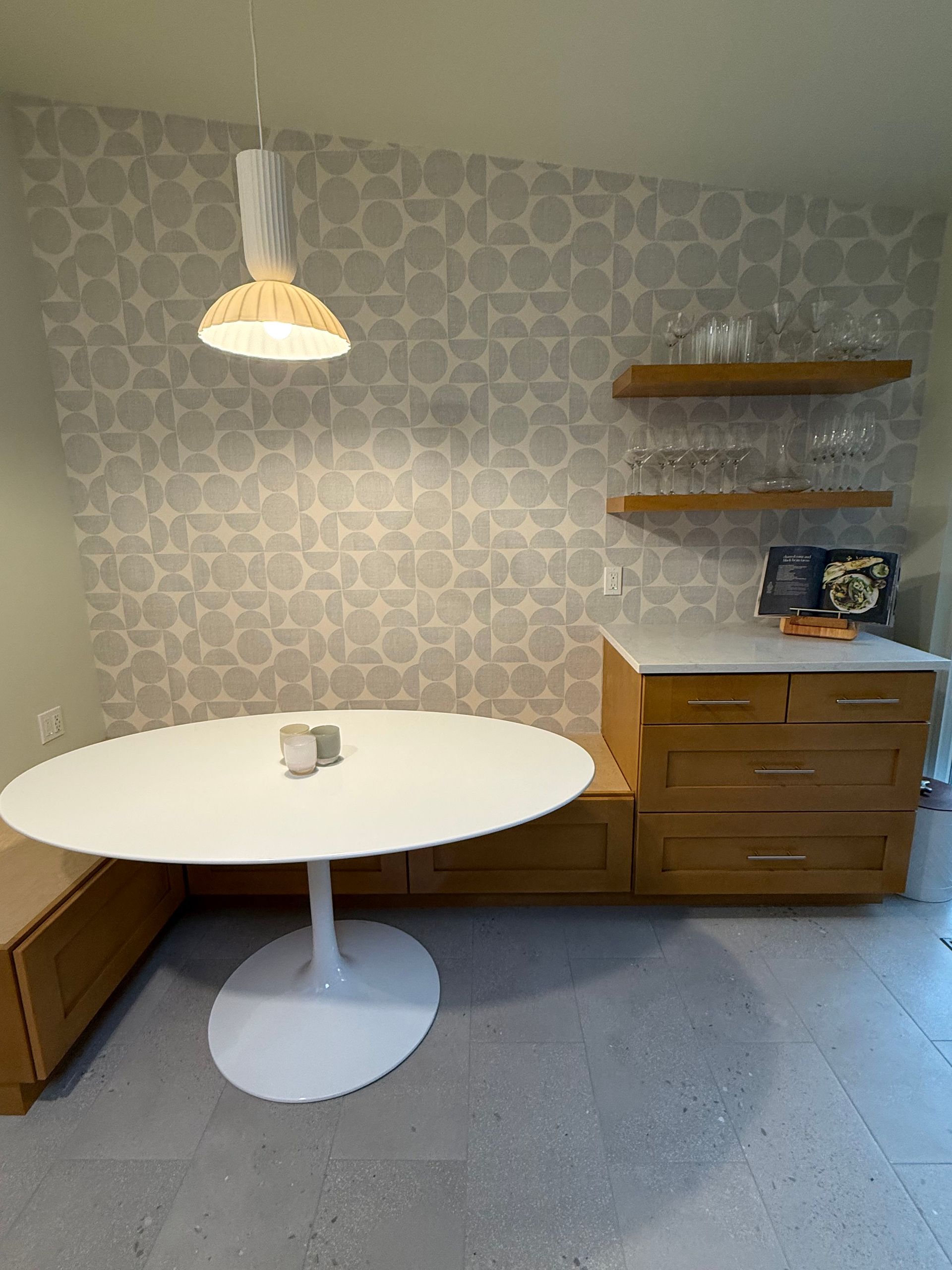 Small dining nook with a white round table, wooden sideboard, wall shelves, and a hanging lamp.