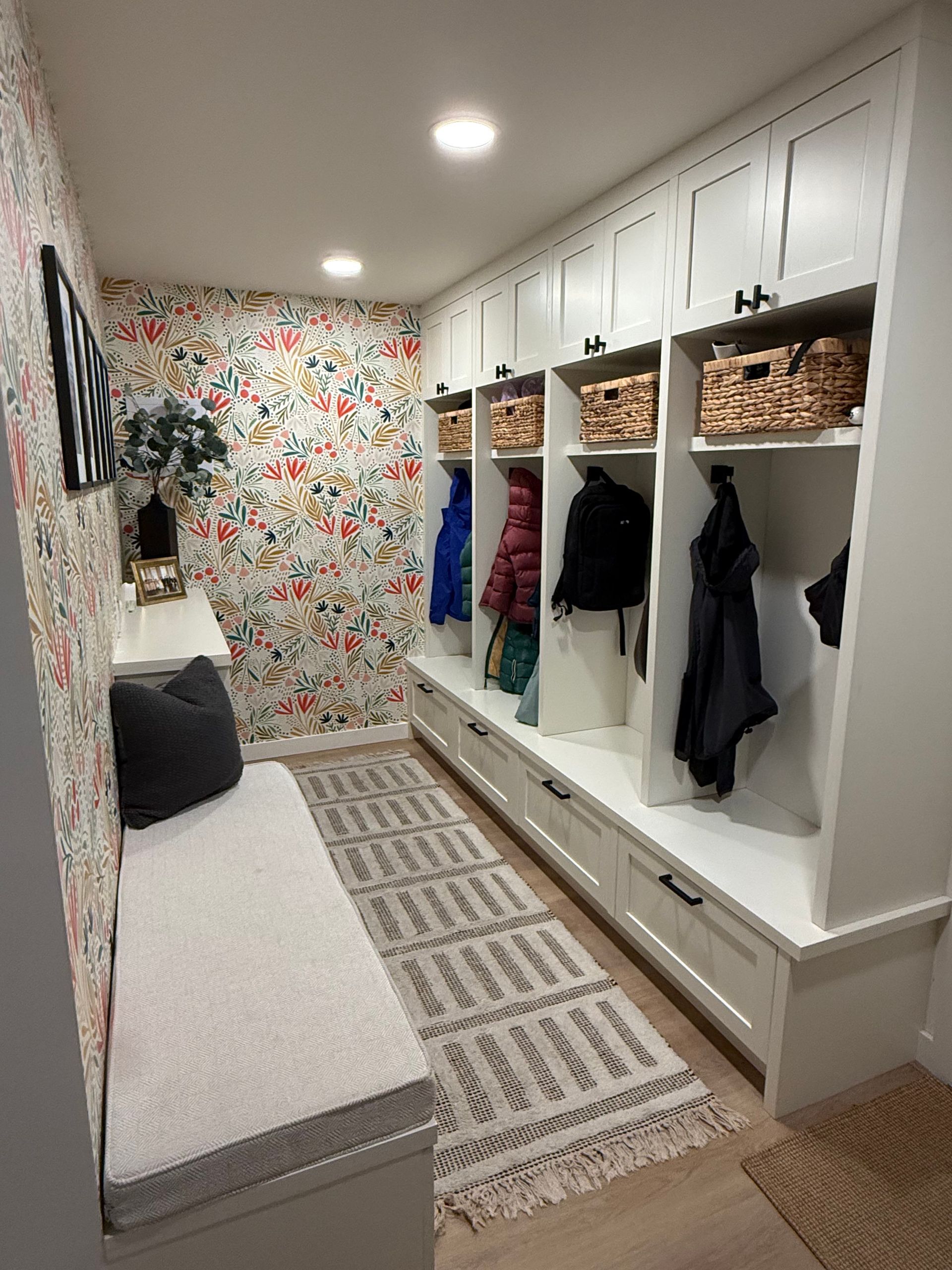 Bright mudroom with white built-in cubbies, baskets, coats, bench, patterned wallpaper, and rugs.