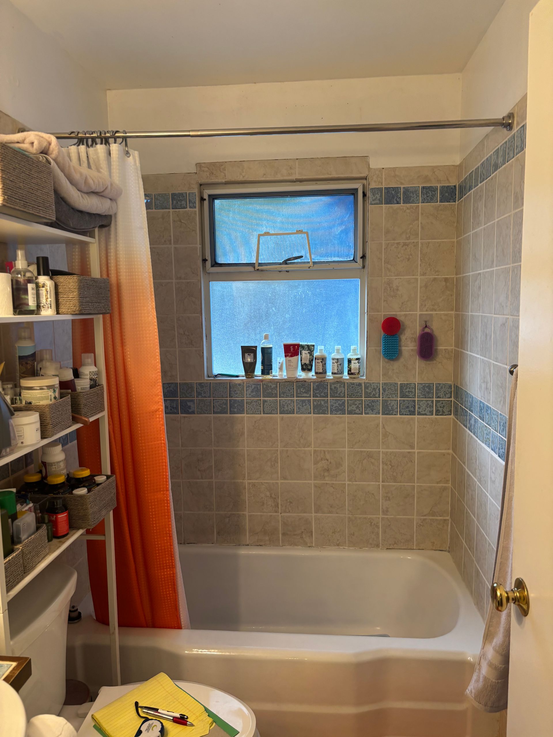 Small bathroom with a tiled bathtub, window, orange shower curtain, and shelves of toiletries