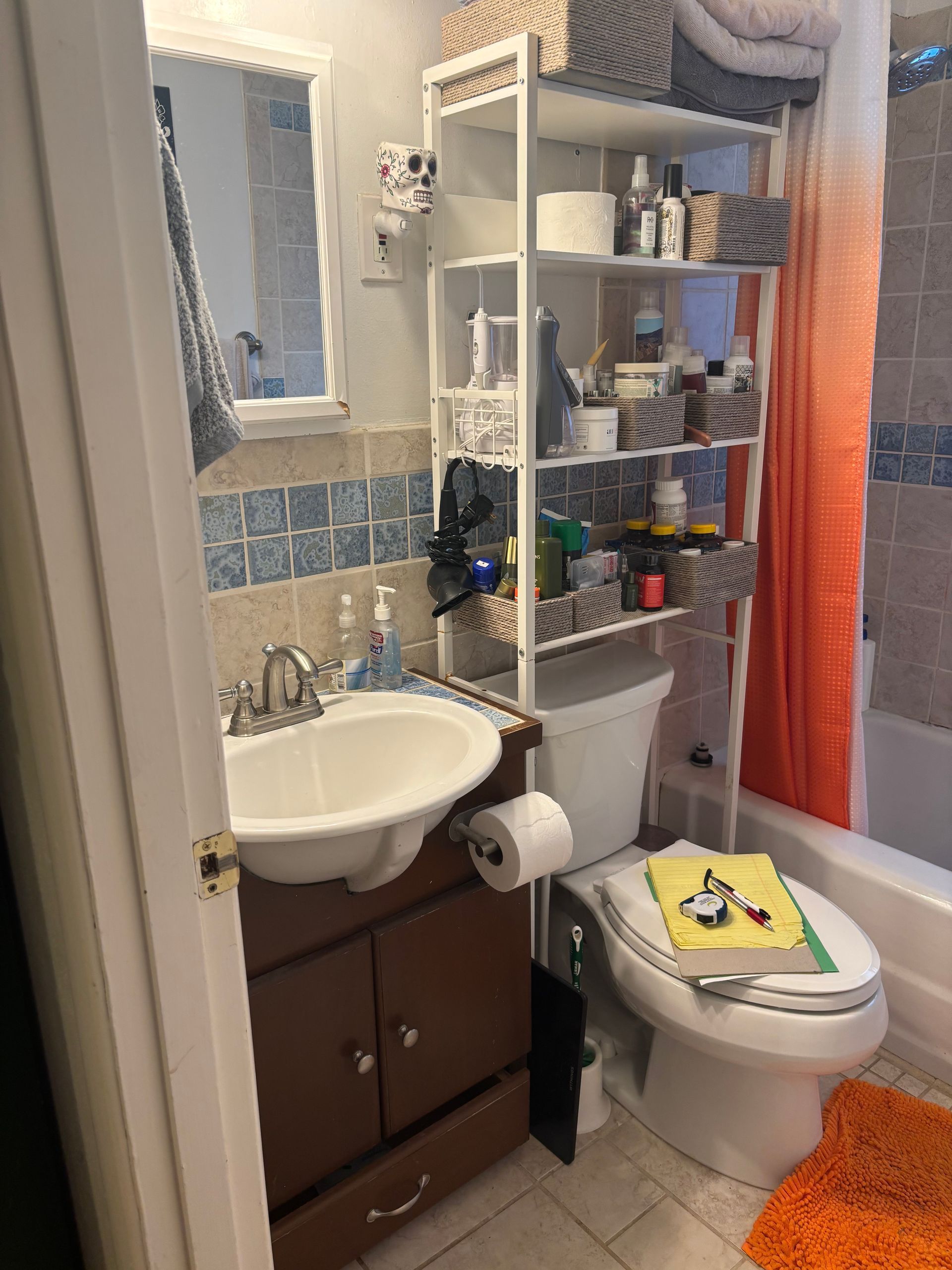 Small bathroom with sink, toilet, shelving, mirror, and orange shower curtain.