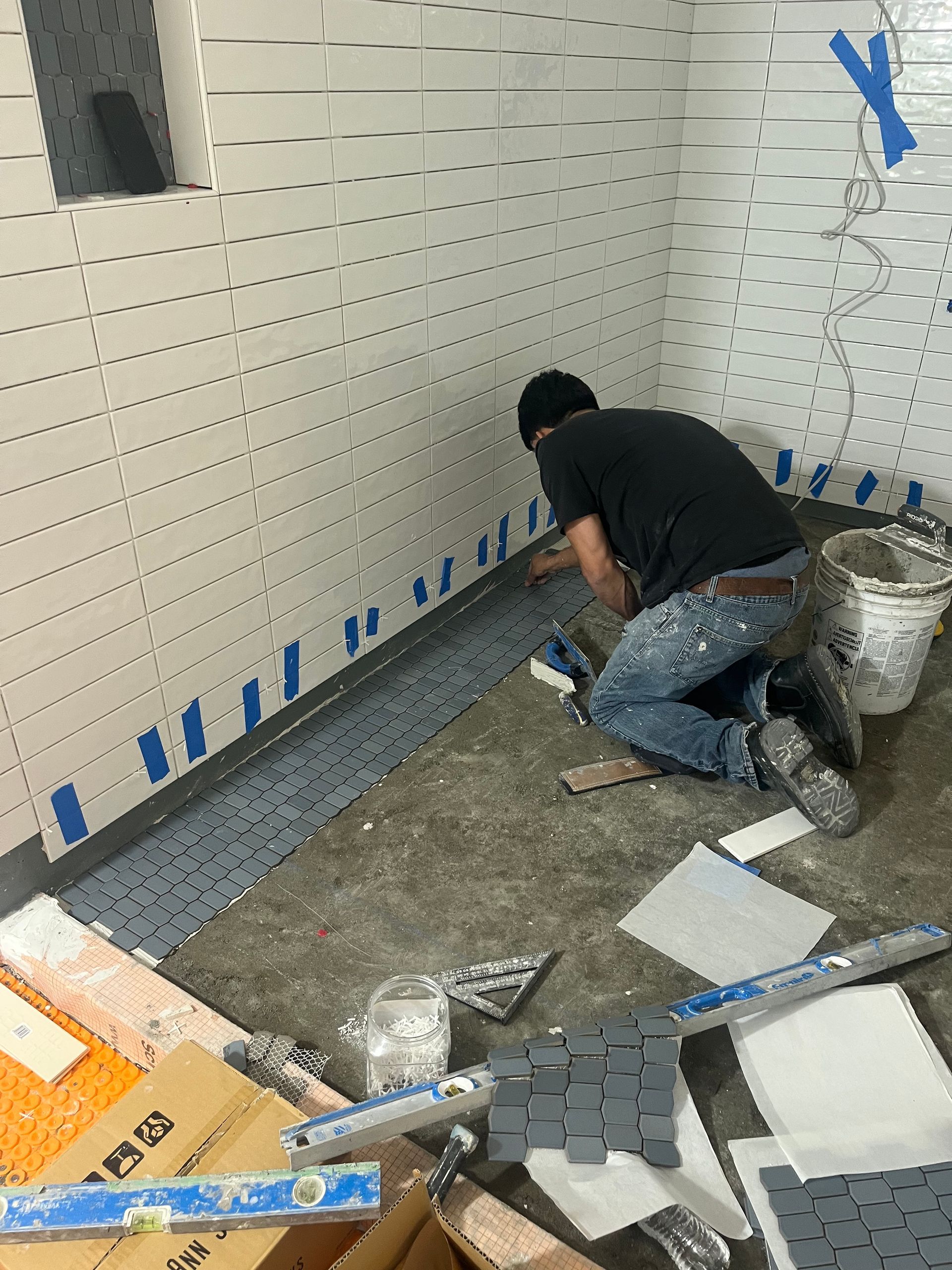 Worker laying gray tile along a wall in a room under construction.
