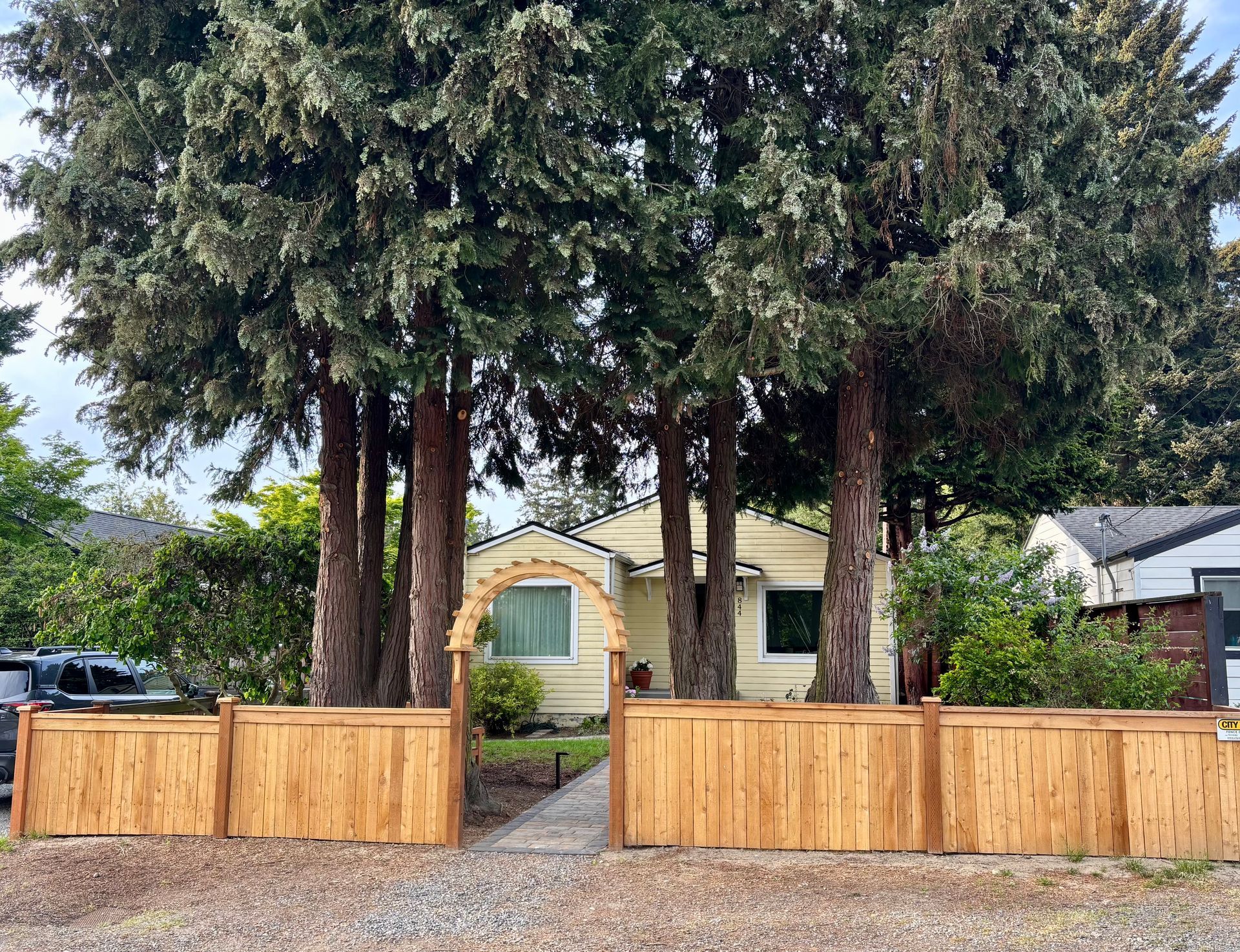 Wooden fence and gate in front of a small house beneath tall evergreen trees.