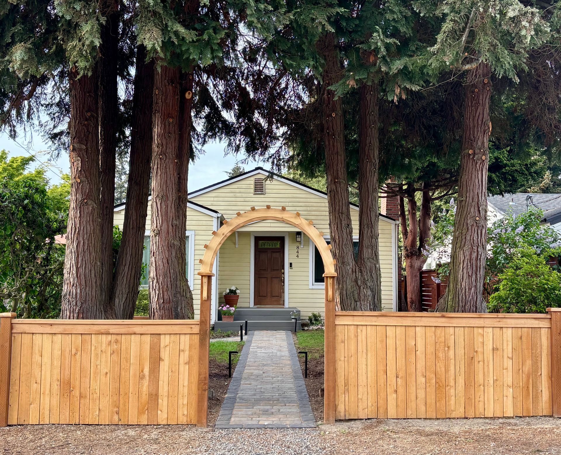 Small house with arched entry behind a wooden fence and tall evergreen trees.