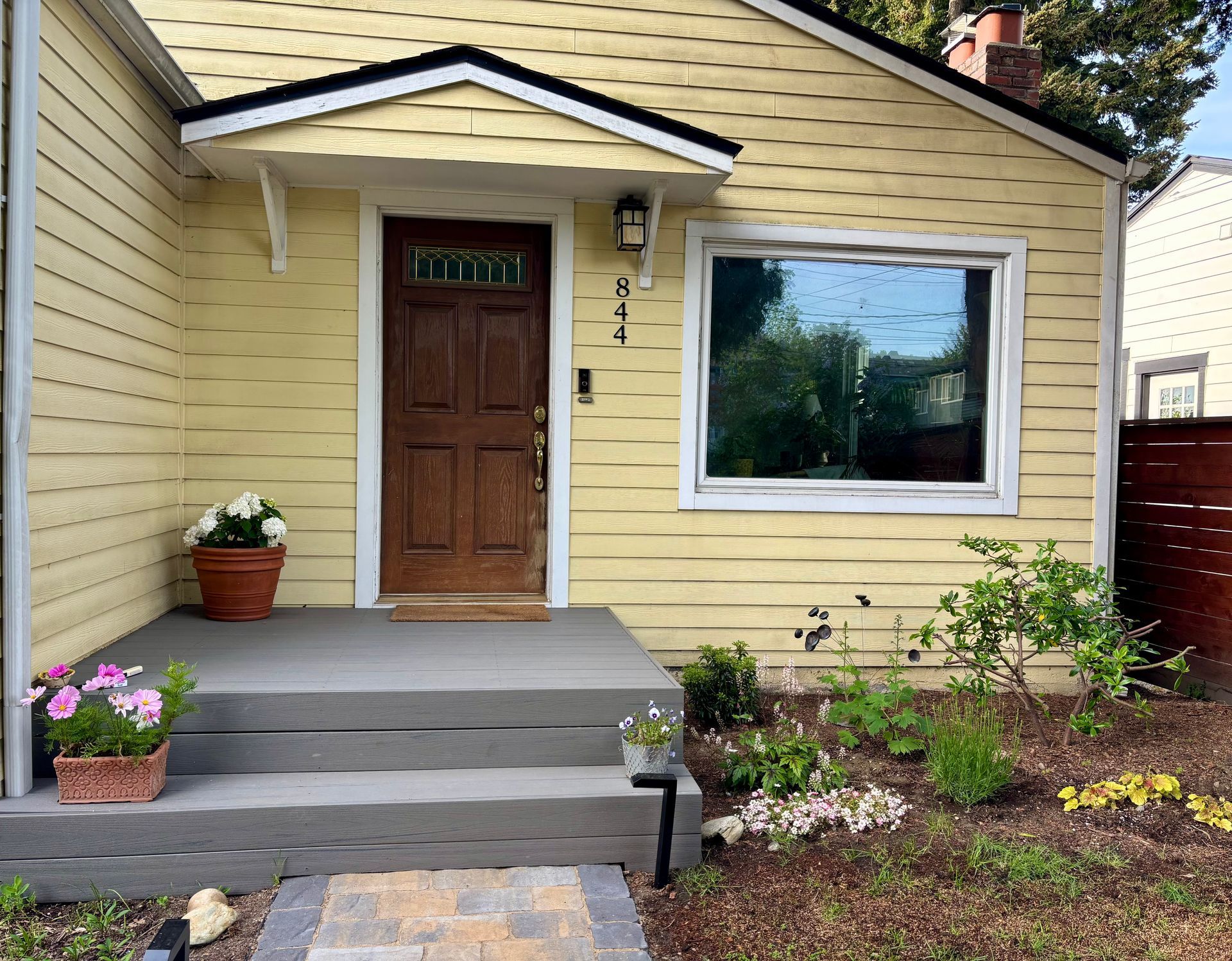 Front porch of a yellow house with a wooden door, large window, and potted flowers.