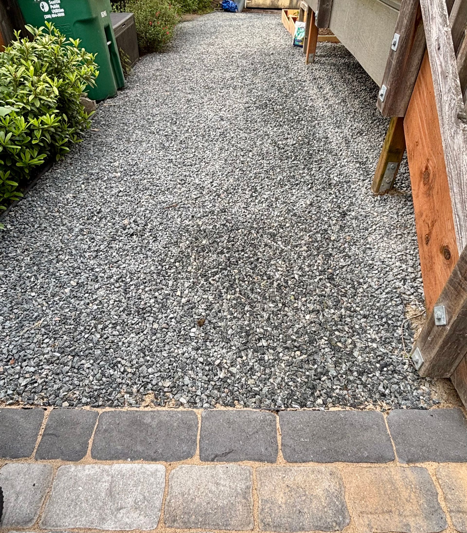 Narrow gravel pathway between a wooden wall and greenery, bordered by stone pavers at the front.