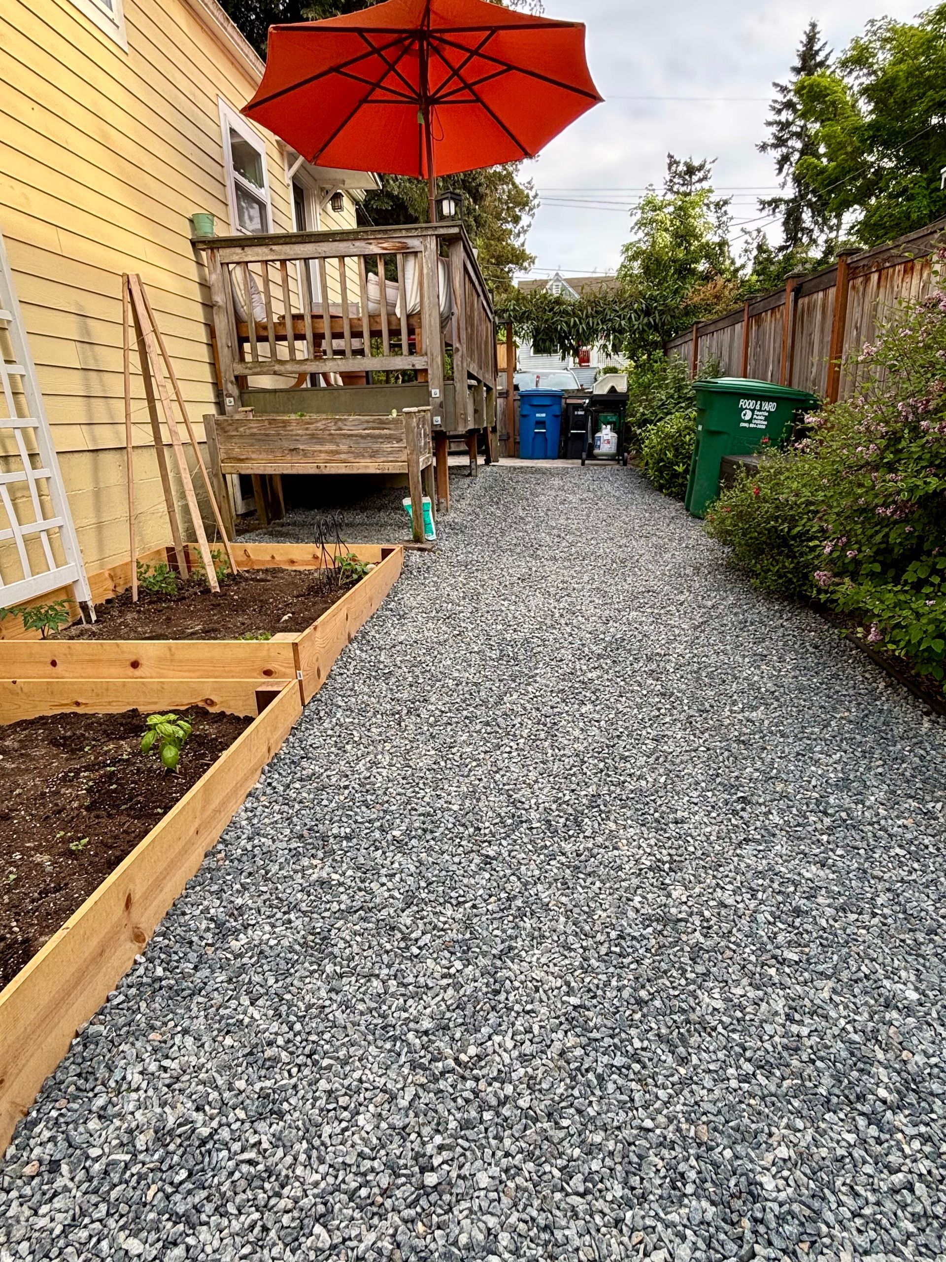 Gravel backyard path beside raised garden beds and a wooden deck with a red umbrella