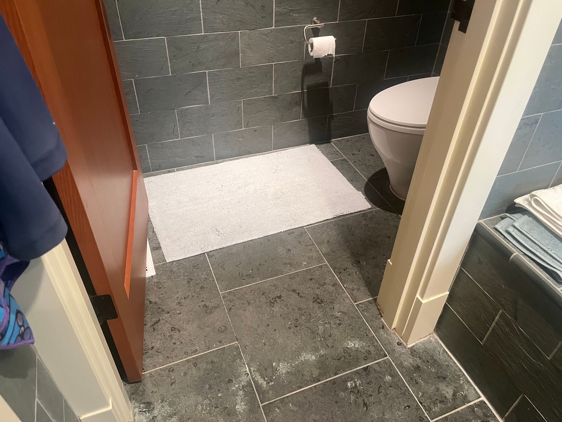 Small bathroom with dark tiled floor, white bath mat, and partially visible toilet and shower doorway.
