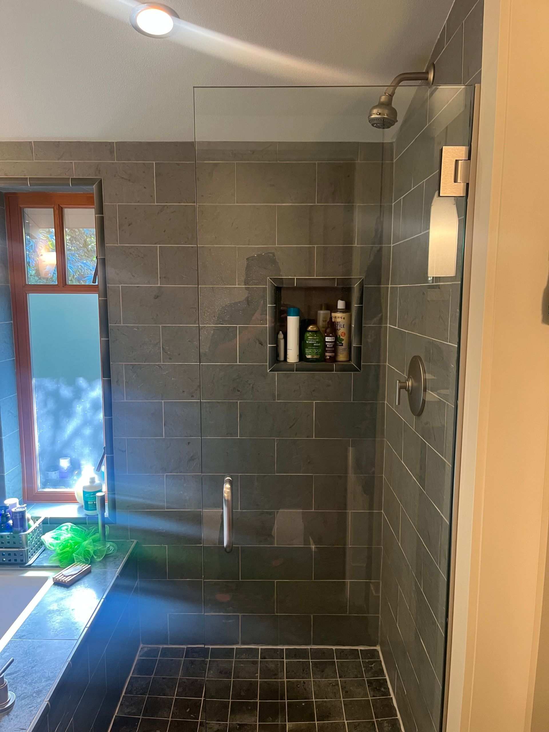 Modern gray-tiled bathroom with a shower, narrow window, and wall shelf of toiletries