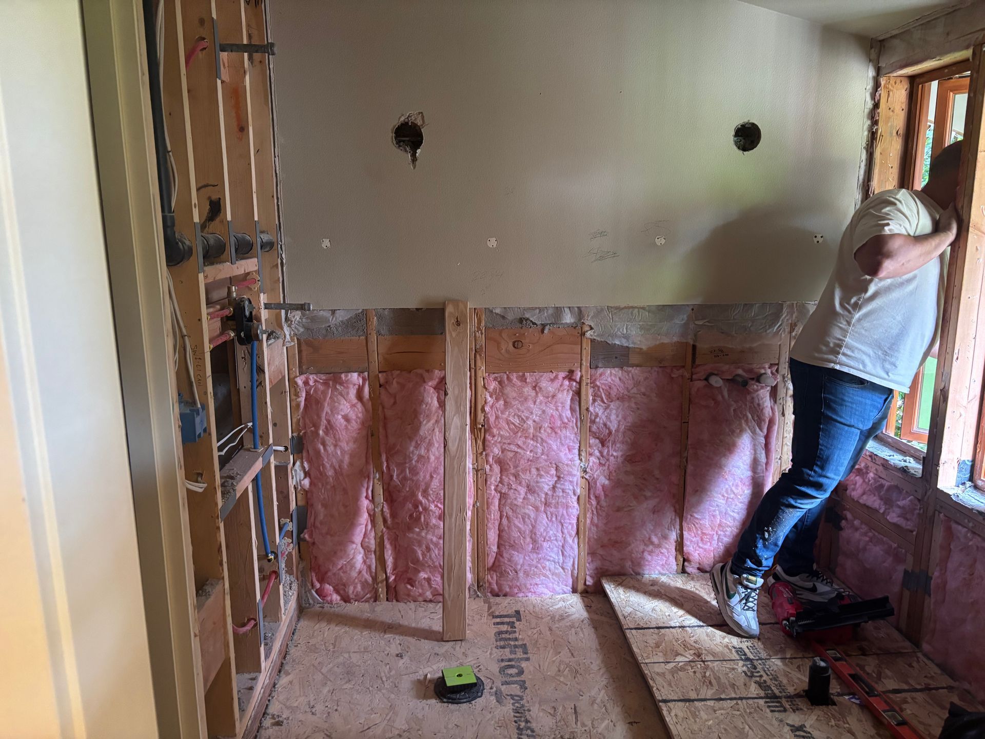 Person standing in a partially insulated room with exposed wall studs and pink insulation, near a window.