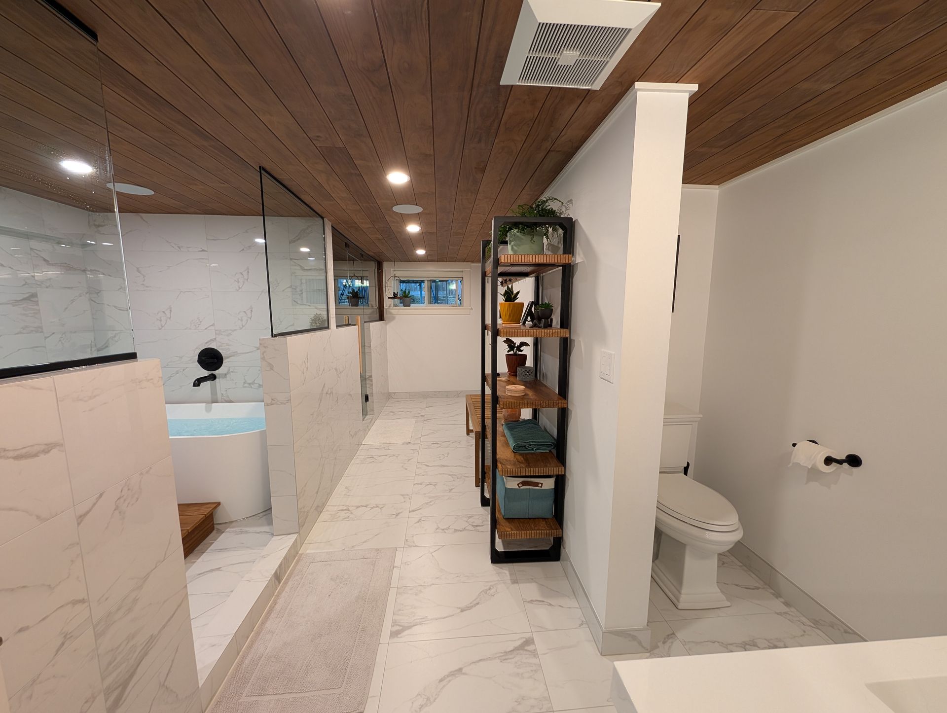 Modern bathroom with marble tile, wood ceiling, glass shower, toilet, and black shelving unit.