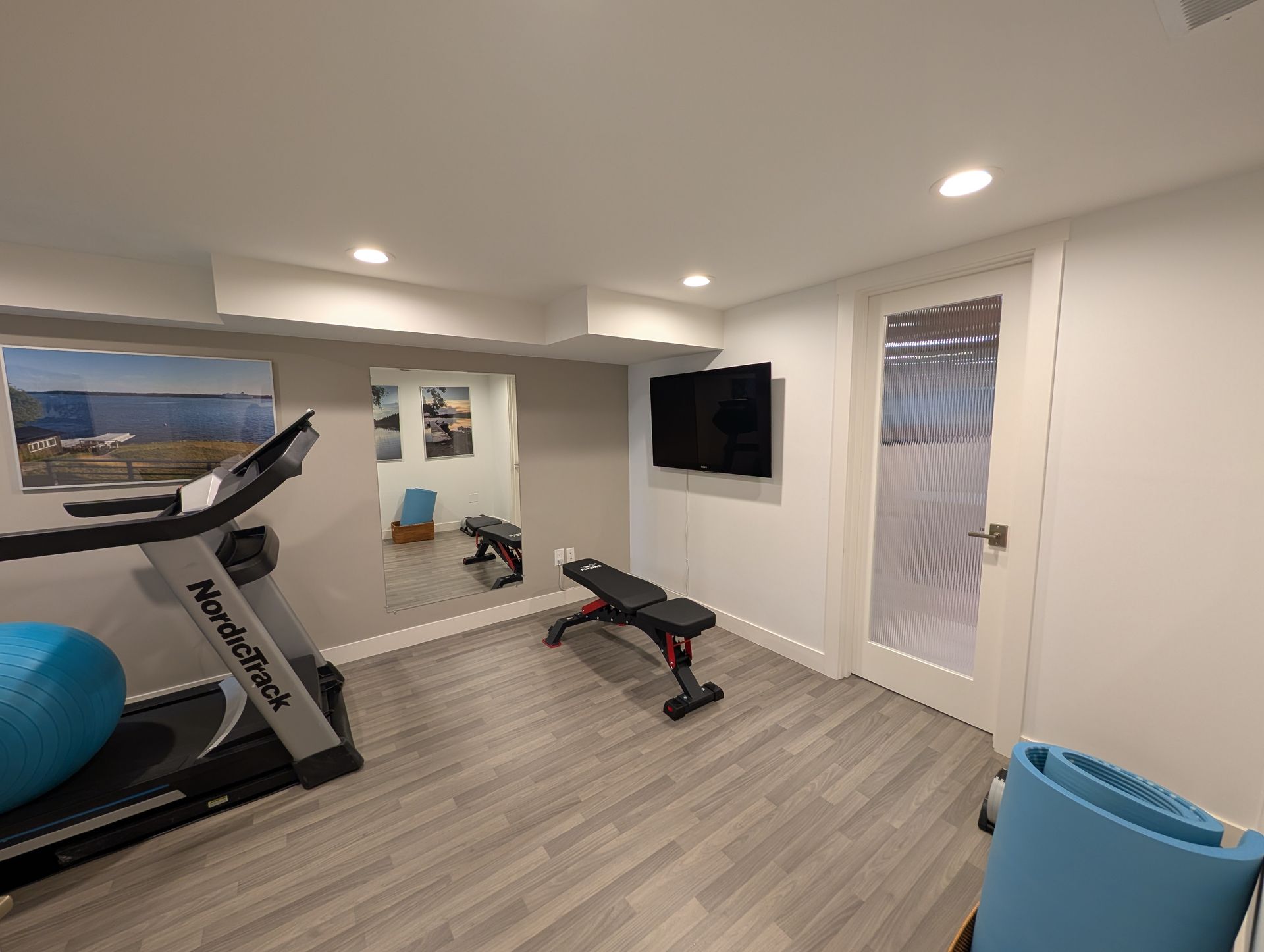Home gym with treadmill, weight bench, TV, blue exercise ball, and glass door in a bright room