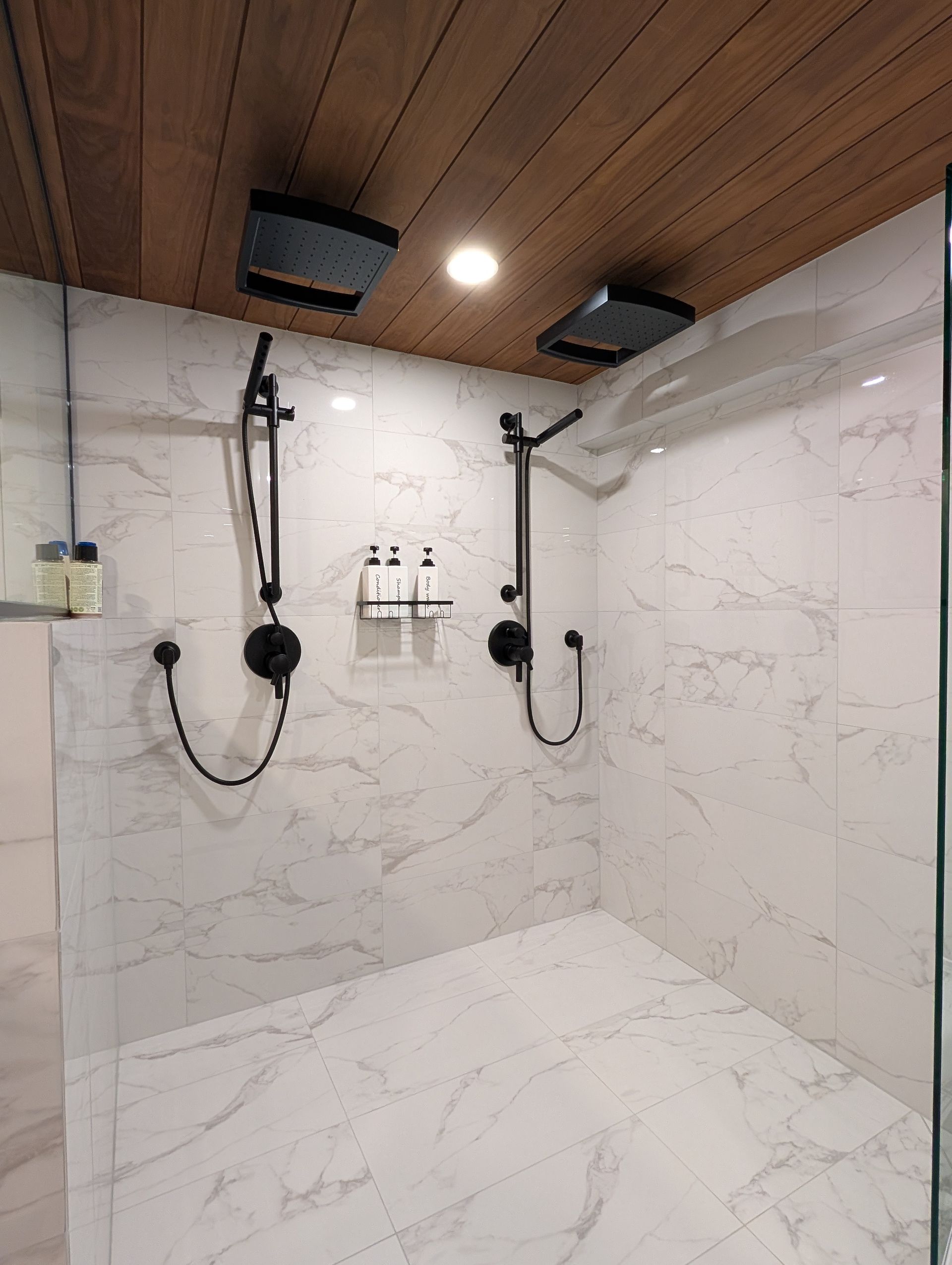Modern shower with marble tile walls, wood ceiling, and two black showerheads with controls