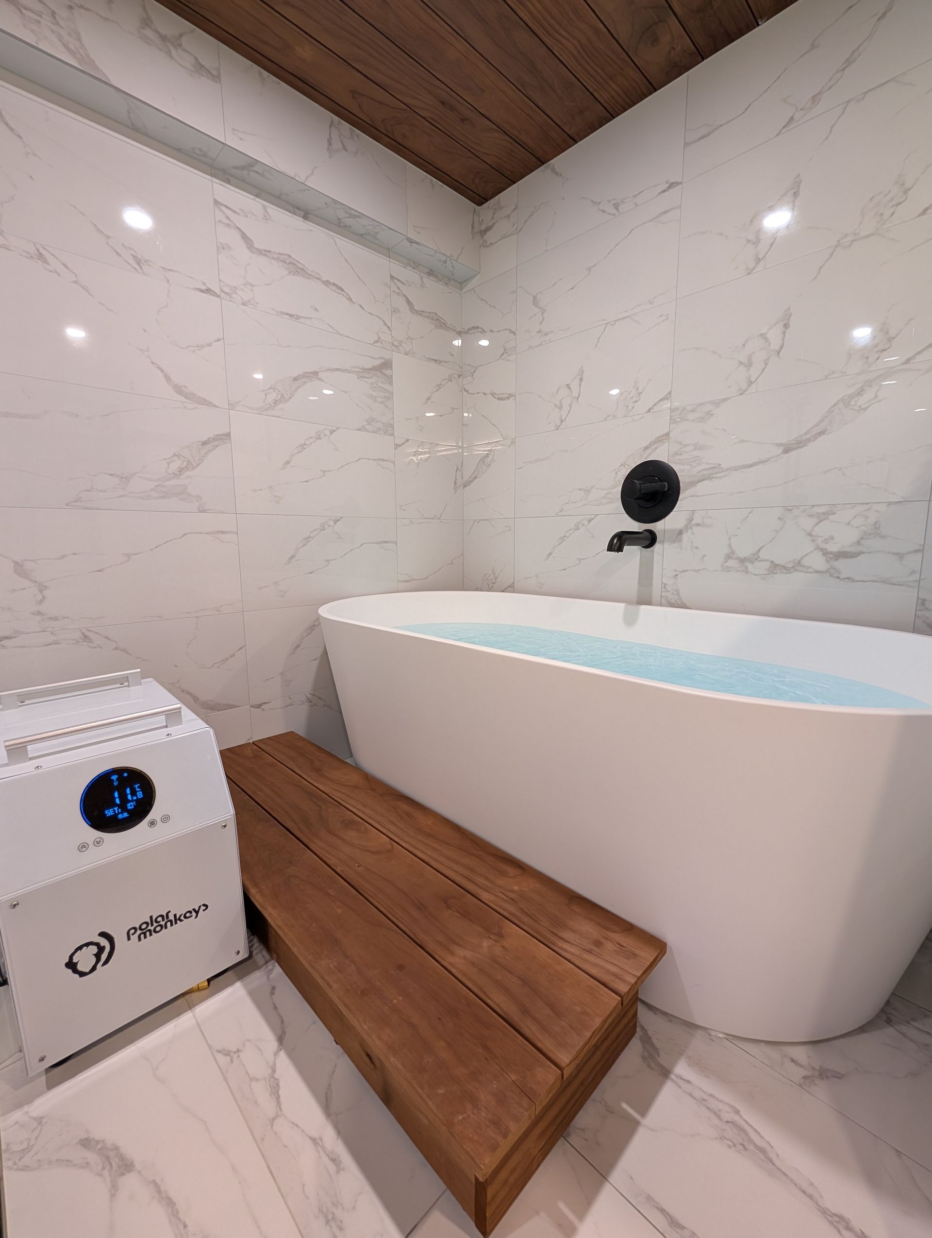 Modern bathroom with marble walls, wood bench, and a freestanding tub under a sloped wooden ceiling