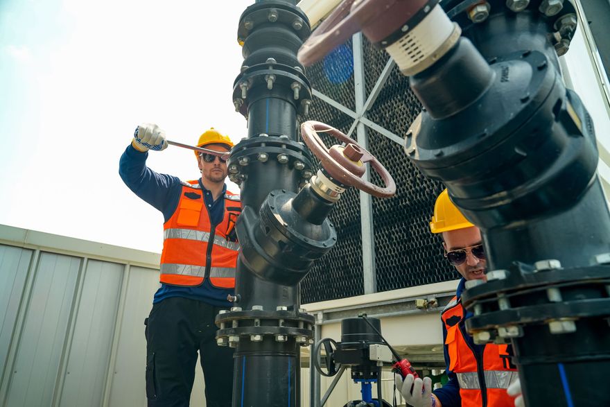 Deux ouvriers portant des gilets de sécurité orange inspectent de grandes canalisations industrielles à l'extérieur.