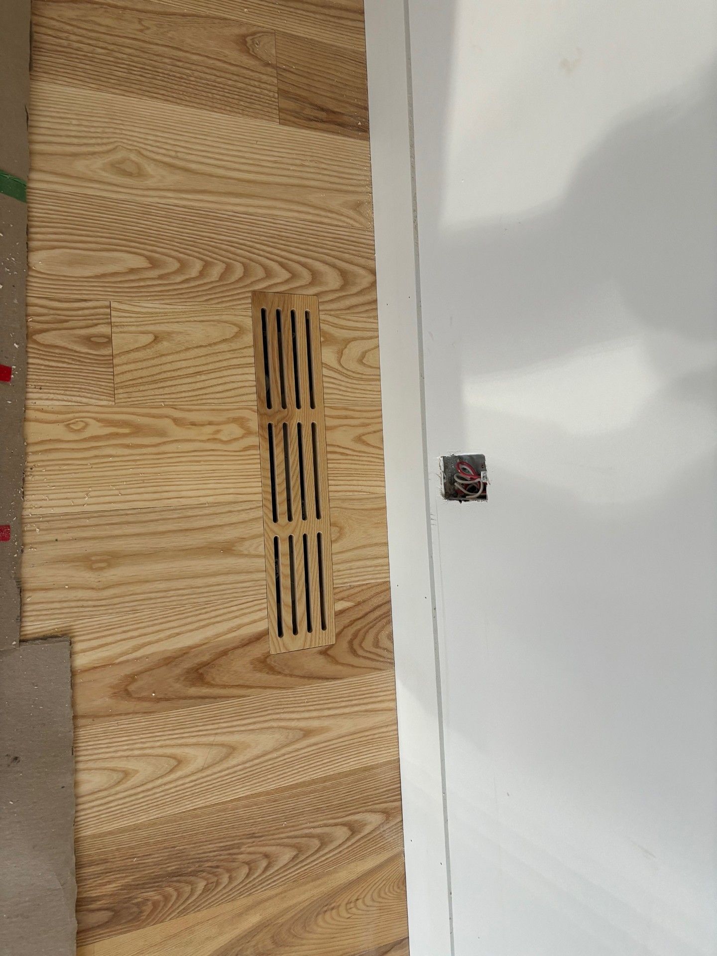 Sol en bois avec une grille d'aération, à côté d'un mur blanc muni d'une prise électrique.